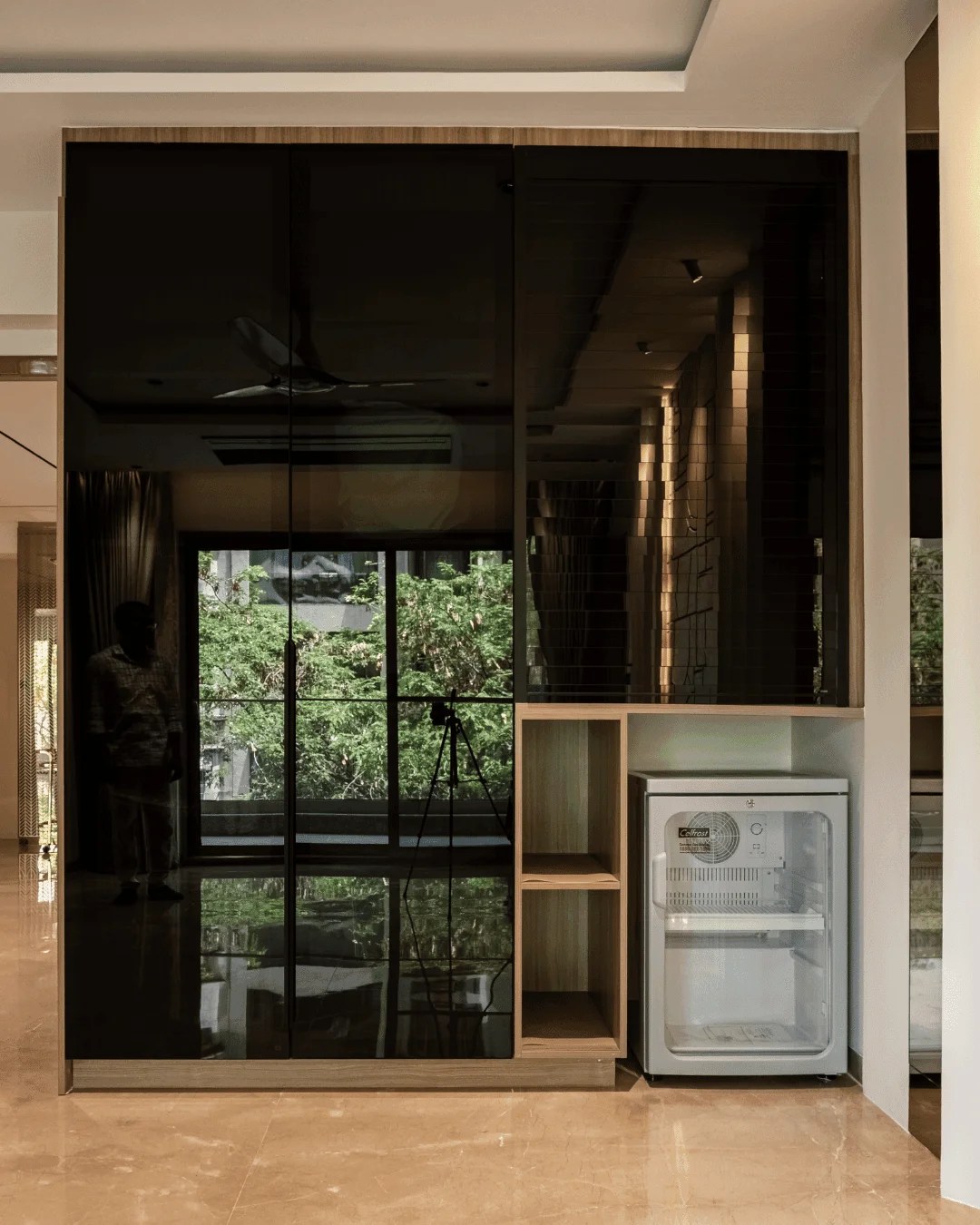 A luxury private residence bedroom featuring a premium tinted glass wardrobe that elegantly reflects the outdoor greenery. The custom unit integrates warm wood framing, sleek open display shelving, and a built-in minibar station on polished marble flooring.