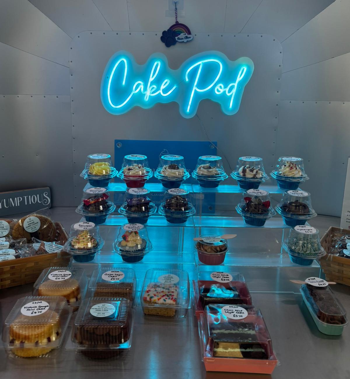 Picture showing a variety of cakes for sale. A blue neon sign spelling "Cake Pod" is located on the rear wall.
