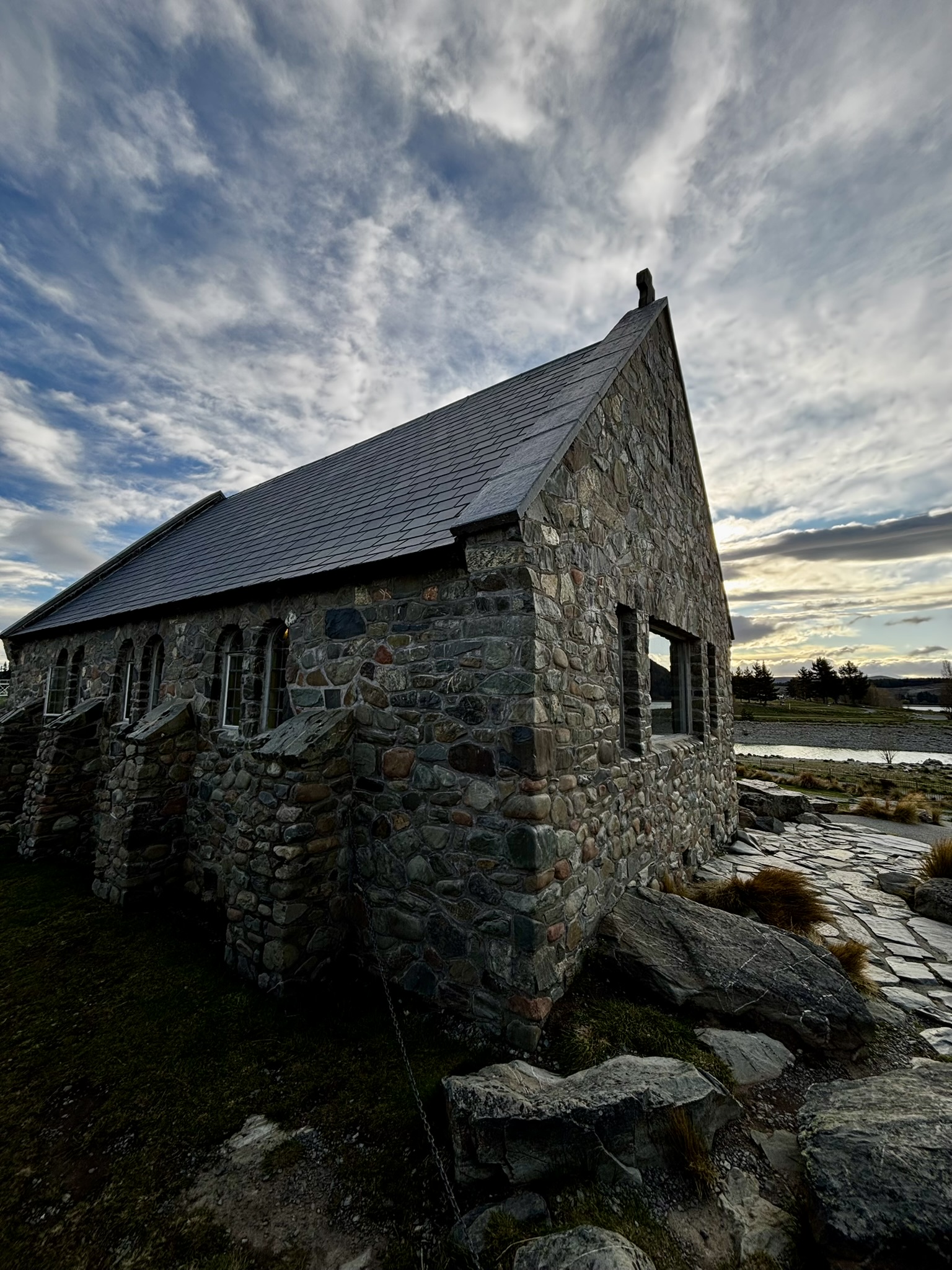 A small stone church with a pointed slate roof is the main feature. The blue sky is fading in twilight and there are atmospheric clouds. The perfect stop during a New Zealand Road Trip.