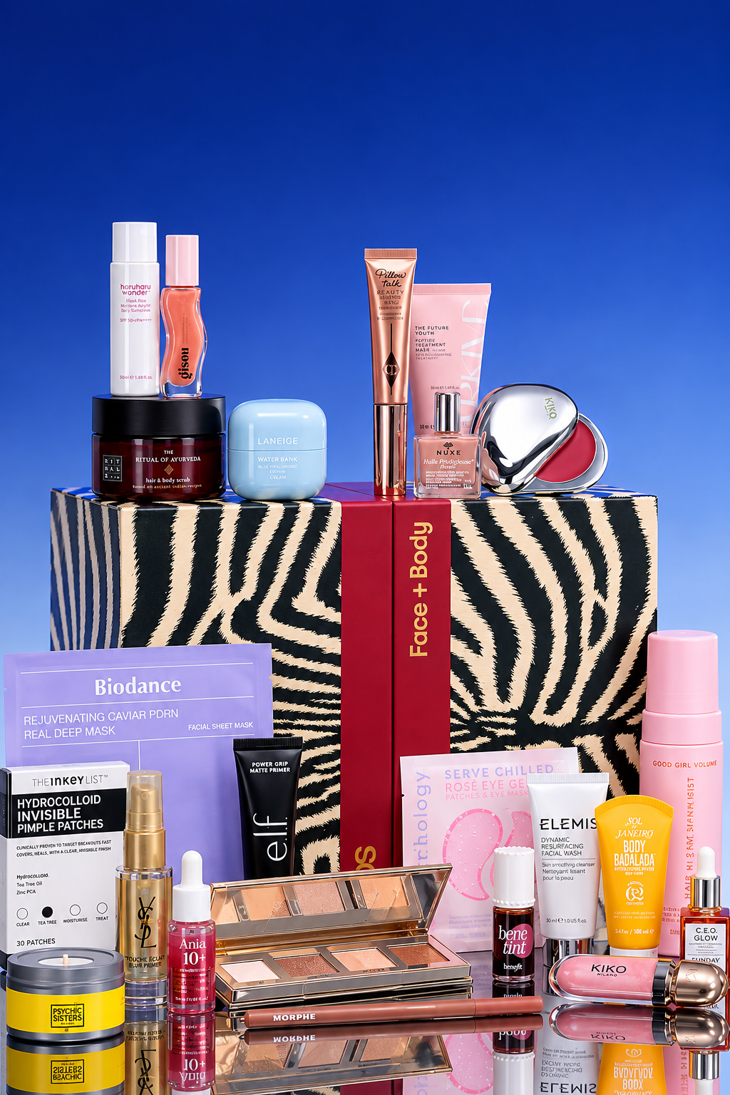 A large array of beauty and skincare products is arranged in front of and on top of a black-and-white zebra-patterned ASOS "Face + Body" box with a vertical red ribbon against a solid blue background. Displayed on top of the box are various items including Haruharu Wonder sunscreen, Gisou hair oil, Rituals hair & body scrub, Laneige Water Bank cream, Charlotte Tilbury Pillow Talk beauty light wand, Nuxe dry oil, and a silver Kiko Milano compact. In front of the box, arranged on a reflective surface, is a selection of skincare, makeup, and hair care items, including a purple Biodance sheet mask, The Inkey List pimple patches, YSL Touche Éclat blur primer, e.l.f. Power Grip primer, a rose gold makeup palette, a Morphe lip pencil, Benefit Benetint, Elemis facial wash, Sol de Janeiro body cream, and a pink spray bottle.