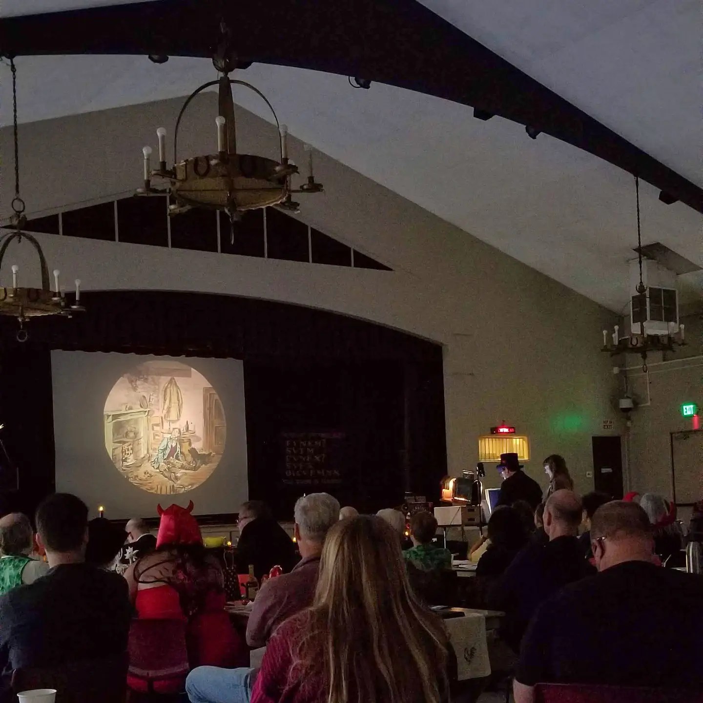 A dimly-lit, large hall with a view of a magic lantern projection on a large screen on the stage, an audience watches.