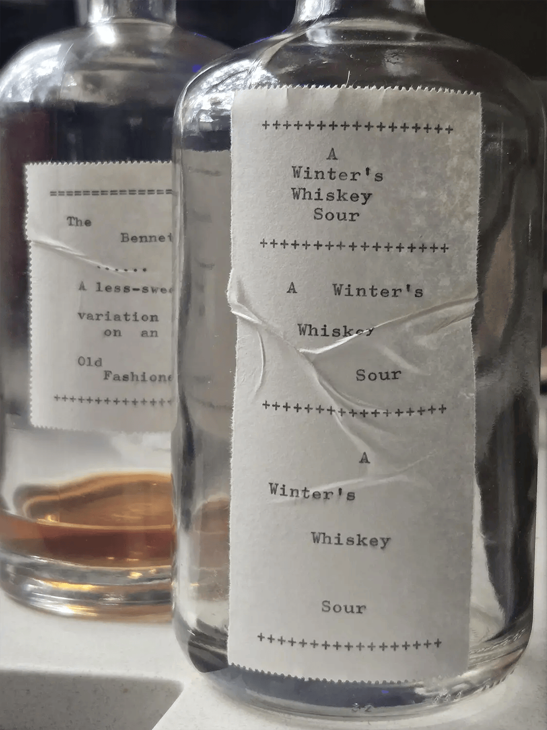 A photo of two, mostly empty cocktail bottles each with a handmade paper label with typewritten text describing the cocktails that the bottles held