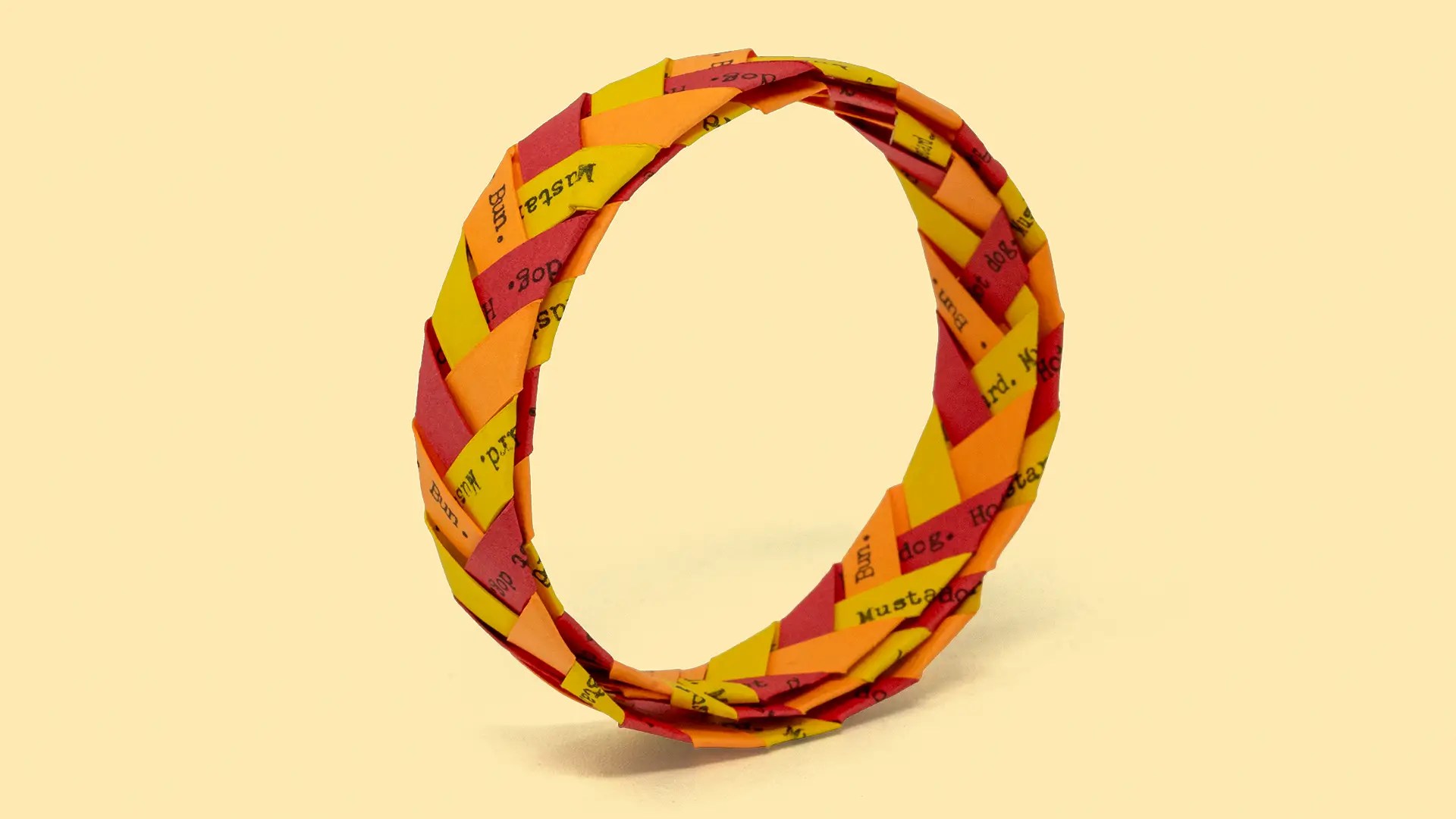 A red, yellow, and orange woven paper bracelet with the words "Mustard," "Hot Dog," and "Bun" repeatedly written in rows by a typewriter.