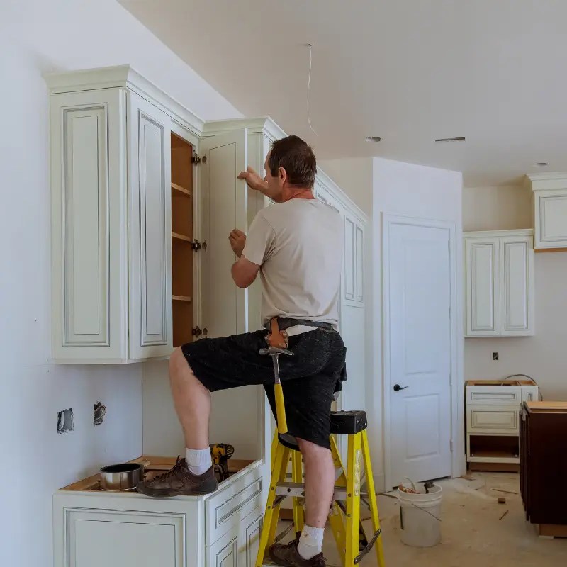 Handyman standing on a step ladder while installing kitchen cabinet doors, holding tools and working in a partially completed kitchen.