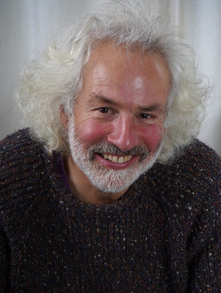 Smiling elderly man with curly gray hair and a beard, wearing a colorful knitted sweater.