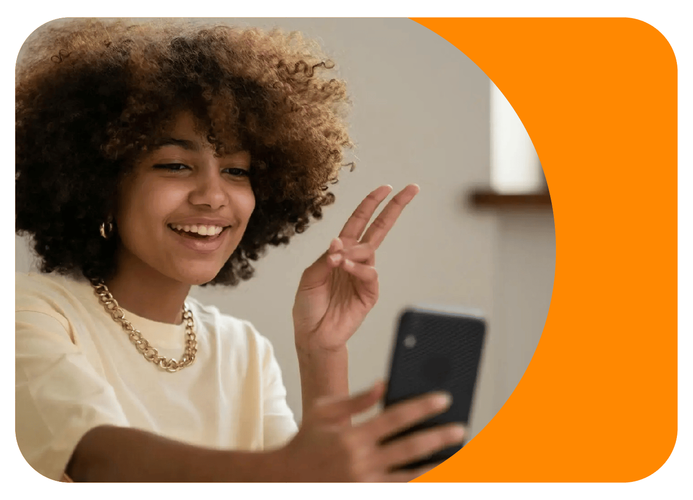 A young woman with curly hair wears a gold chain and is smiling. She's holding a phone with one hand and has a peace sign in the other.