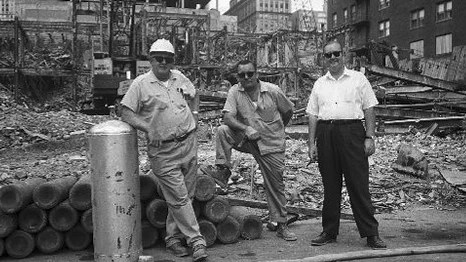 Three men in period clothing stand beside a stack of demolition debris at a historic Hawthorne job site.