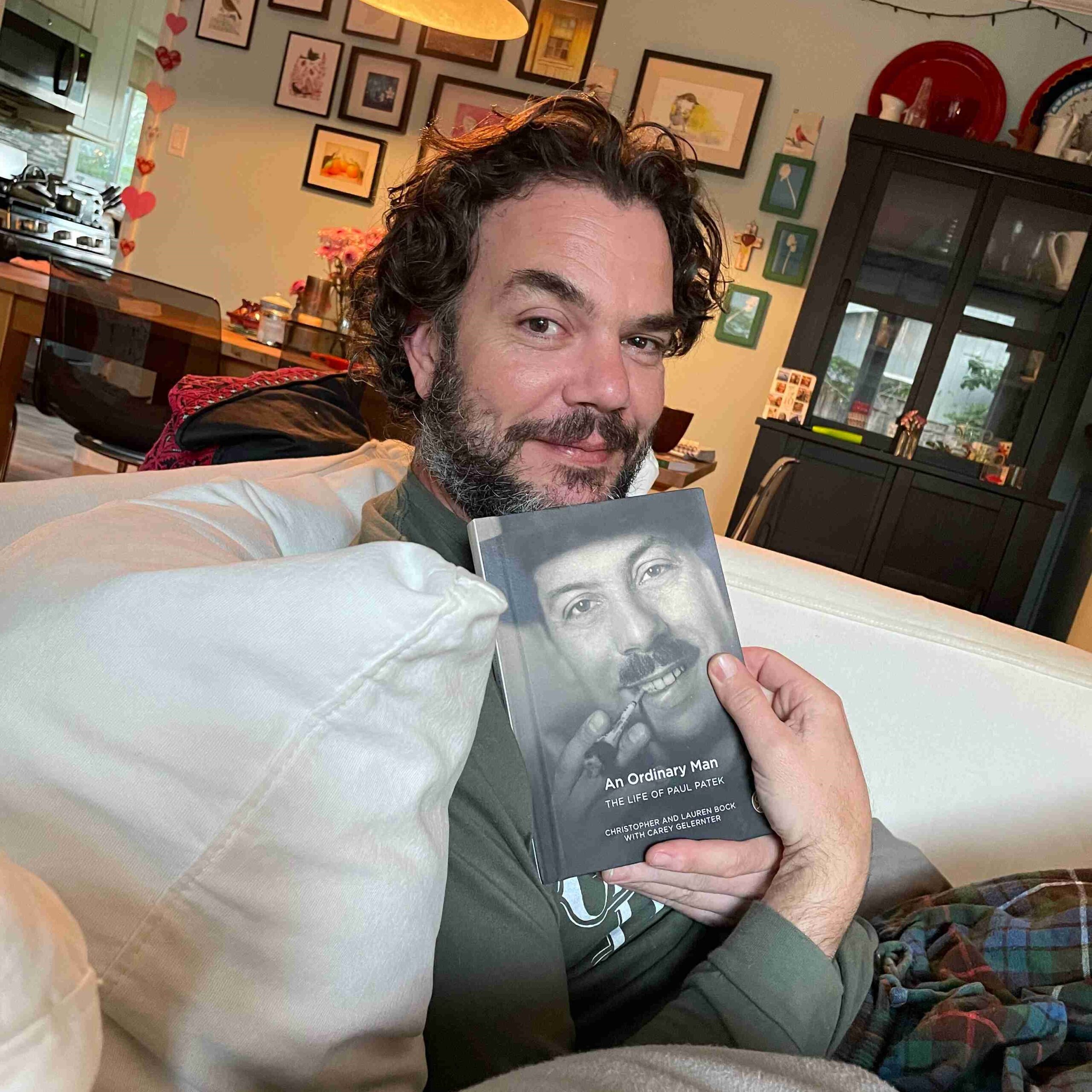 Christopher Bock sitting on a couch showing the cover of his book No Ordinary Man