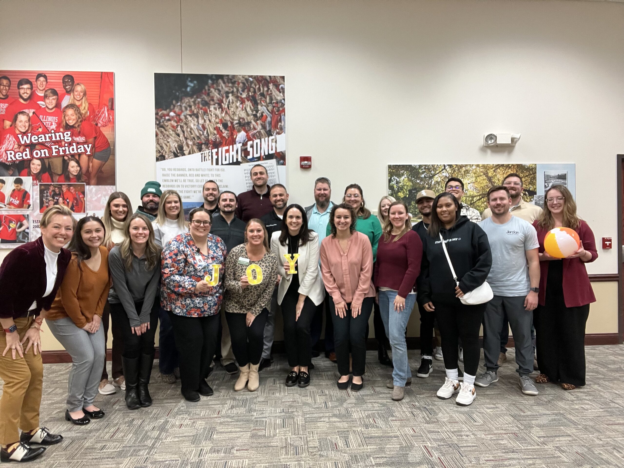 Large group of students and staff posing for a photo in a hallway with posters that say 'Wearing Red on Friday' and 'The Fight Song' in the background, many wearing red.