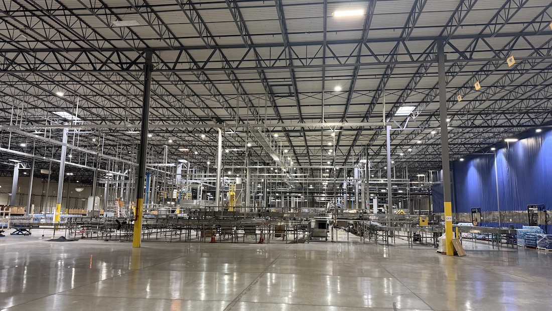 Wide view of a large industrial facility with overhead steel roof trusses, production equipment, and conveyor lines.