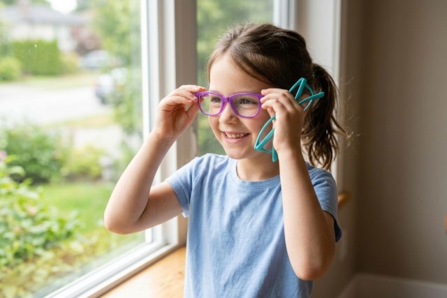 Smiling child trying on purple glasses while holding teal glasses, standing by a window.