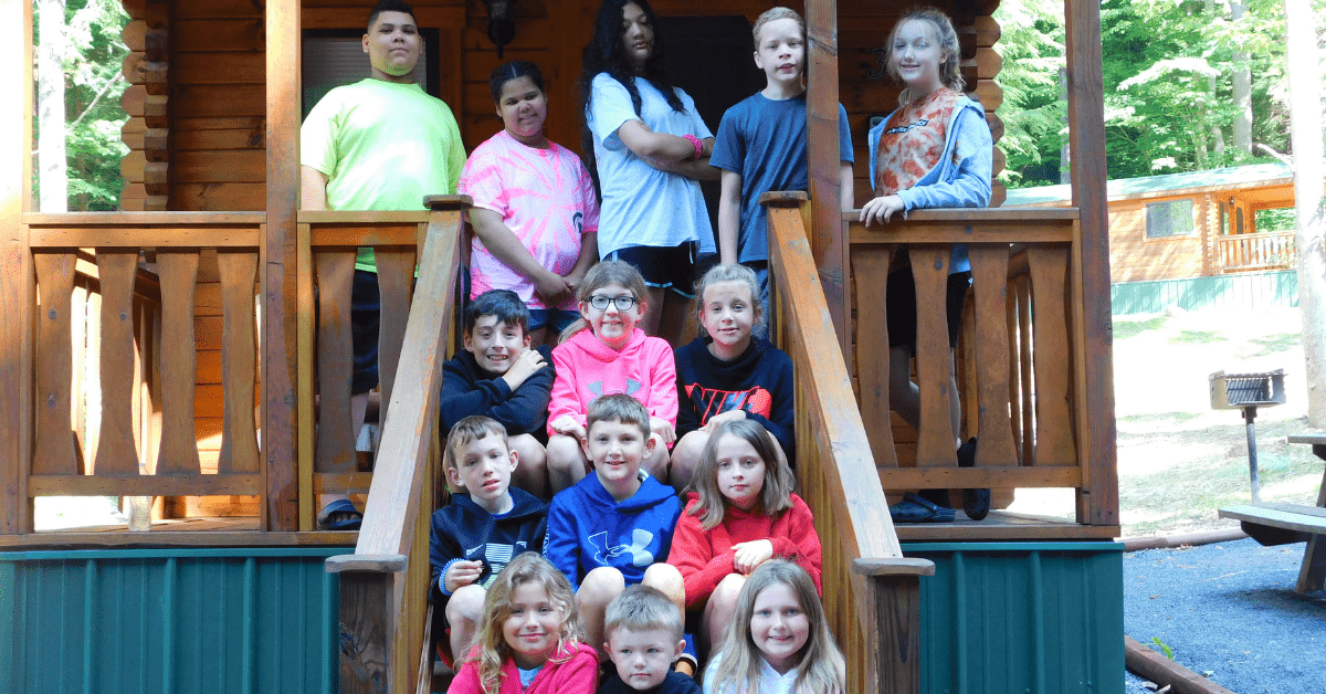 A group of children and teens gathered on the steps of a cabin at a summer camp.