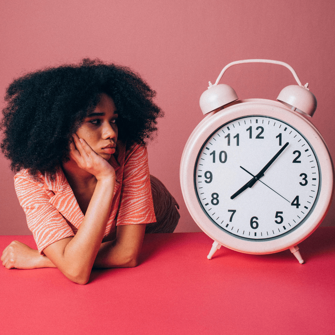 Woman of color lying on her tummy and sitting up on her elbows. She is holding her chin in her right hand as she sadly look at a giant alarm clock right next to her. She has curly hair and the background is a magnified and light pink