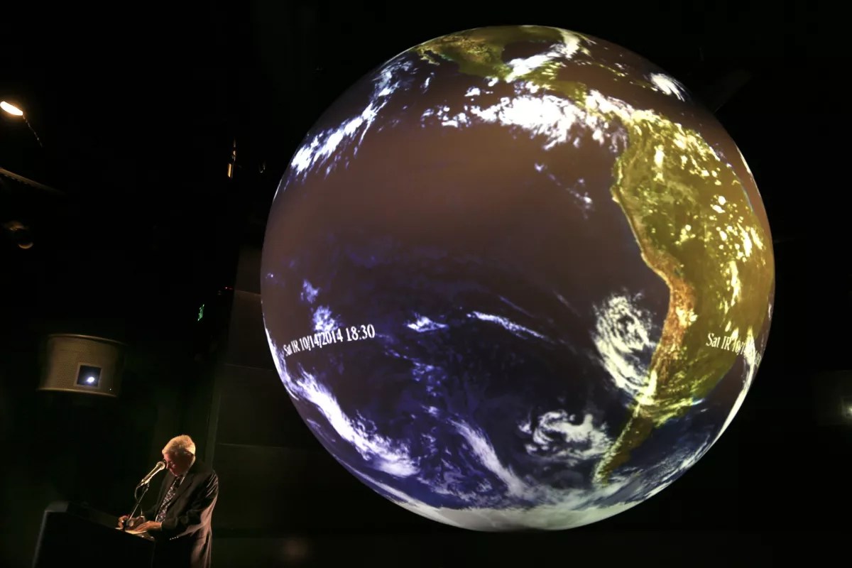 Large sphere on a stage with the features of earth projected on to it. There is a man at a podium to the left of the sphere speaking to a crowd.