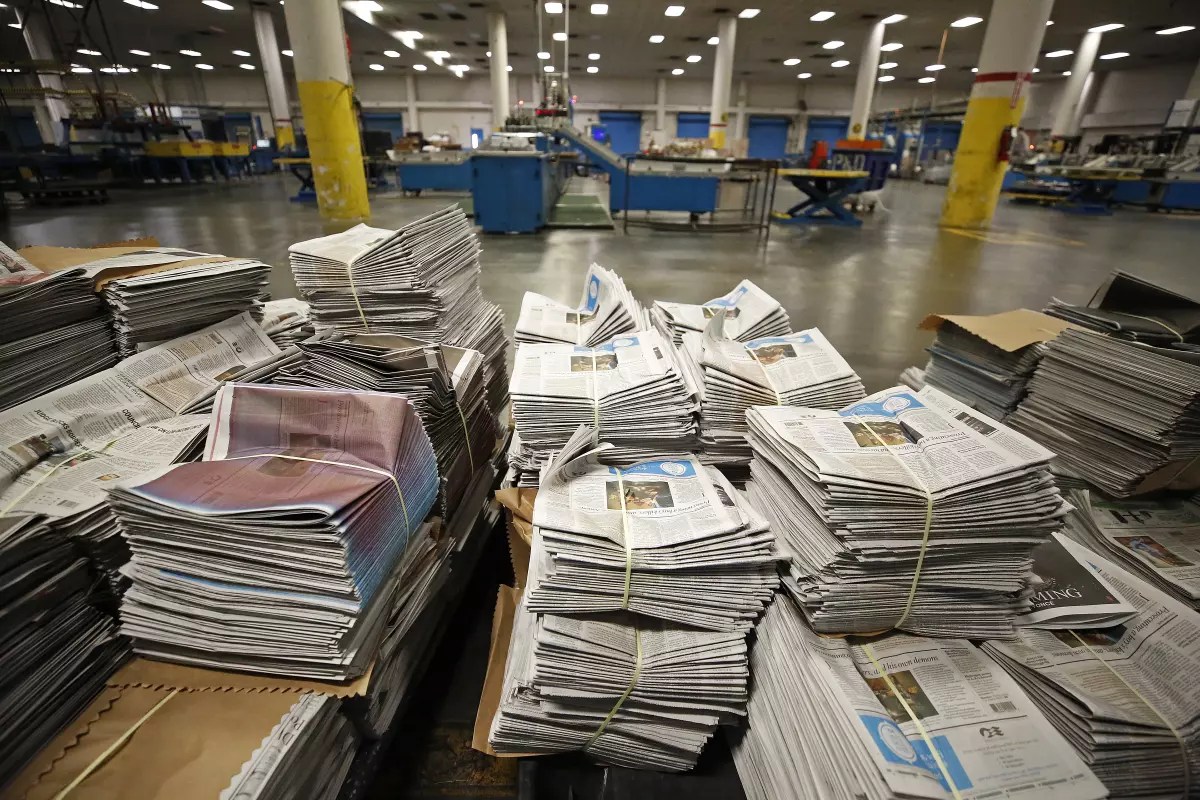Stacks of newspapers sit ready for distribution at The Times’ printing facility south of downtown Los Angeles in 2019. There are about 15 stacks of 100 newspapers in front of a warehouse background.