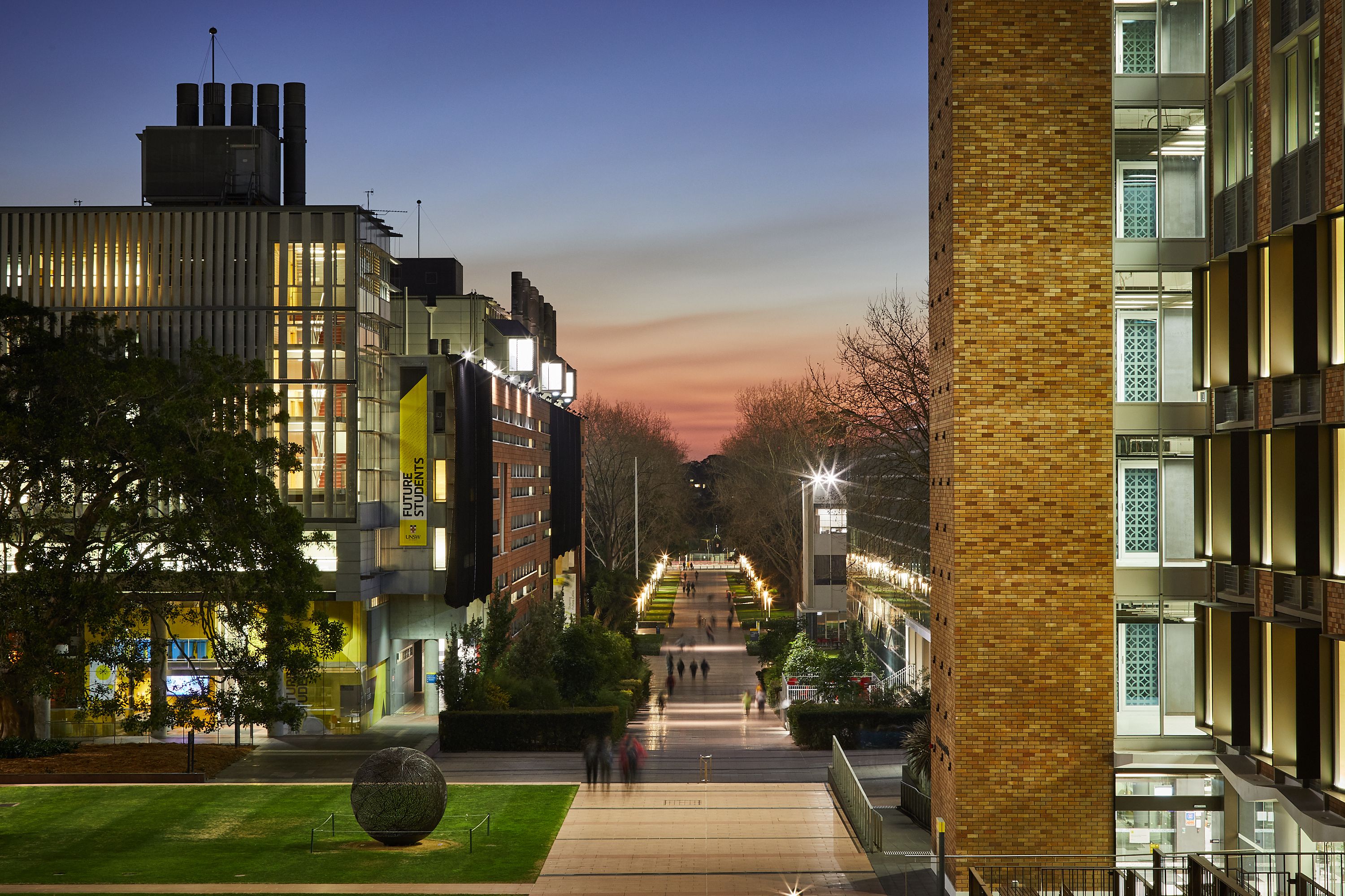 An image of UNSW at night, showing a central walkway between two buildings, with people walking down it.