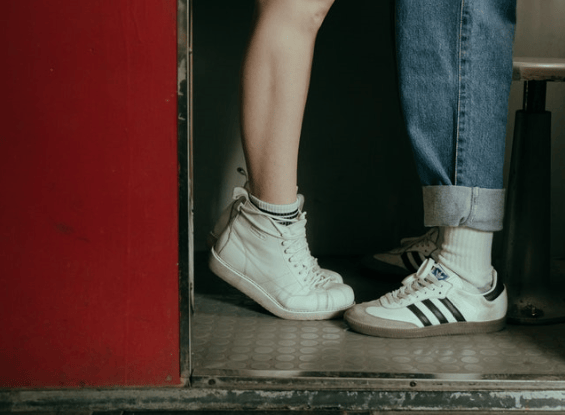 Fotografía de las piernas de una pareja de adolescentes. Se encuentra uno frente al otro. Sólo se visualiza de la rodilla para abajo. Ella se encuentra de puntitas por lo que pareciera que da un beso.