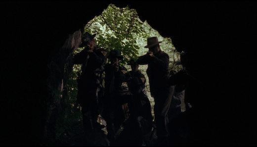 A group of policemen crowd the mouth of a cave, guns pointed inward.