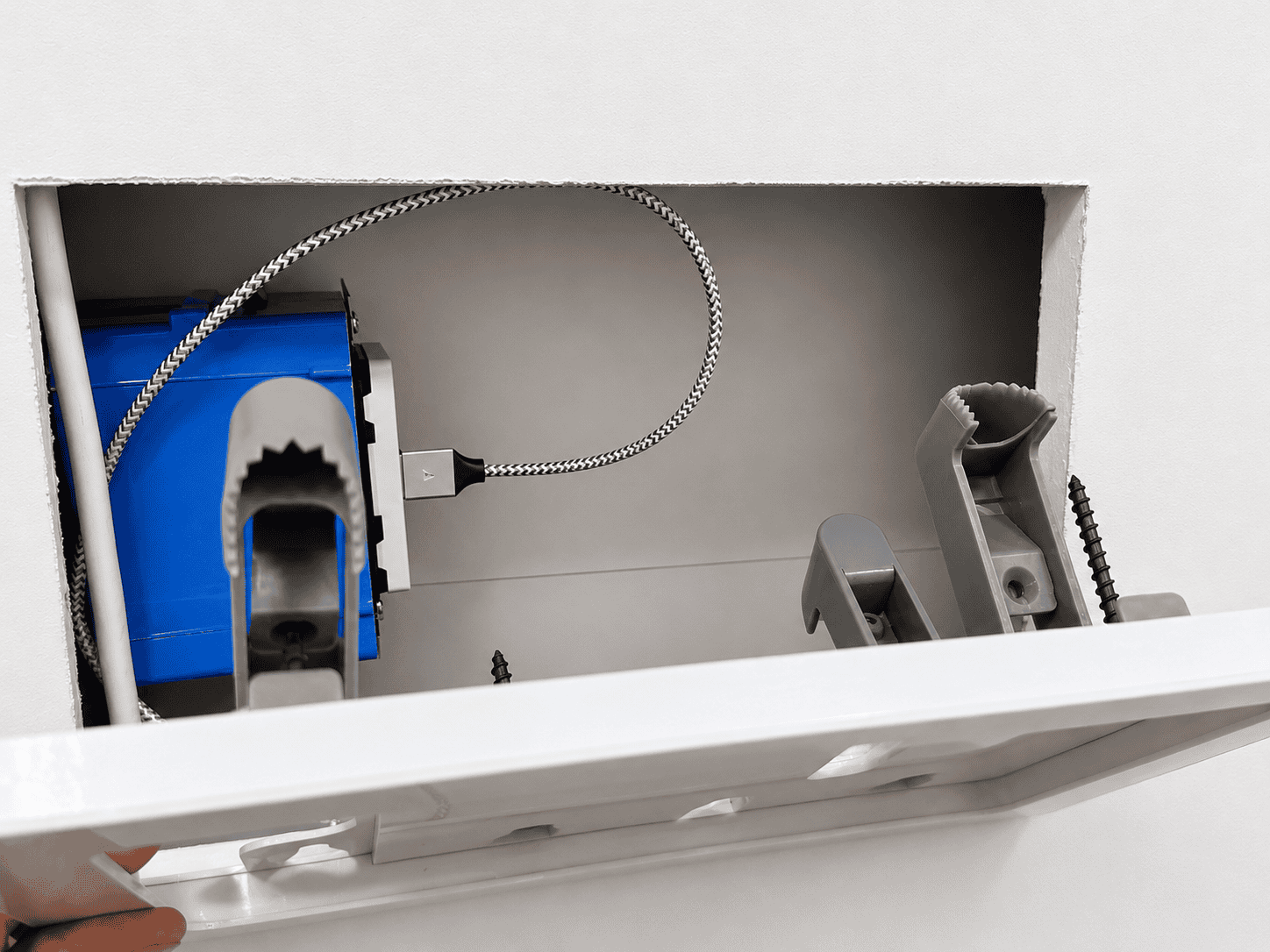 Close-up view of an electrical box with a blue component and a braided cable, set in a wall opening, showing attachment clips and mounting screws.