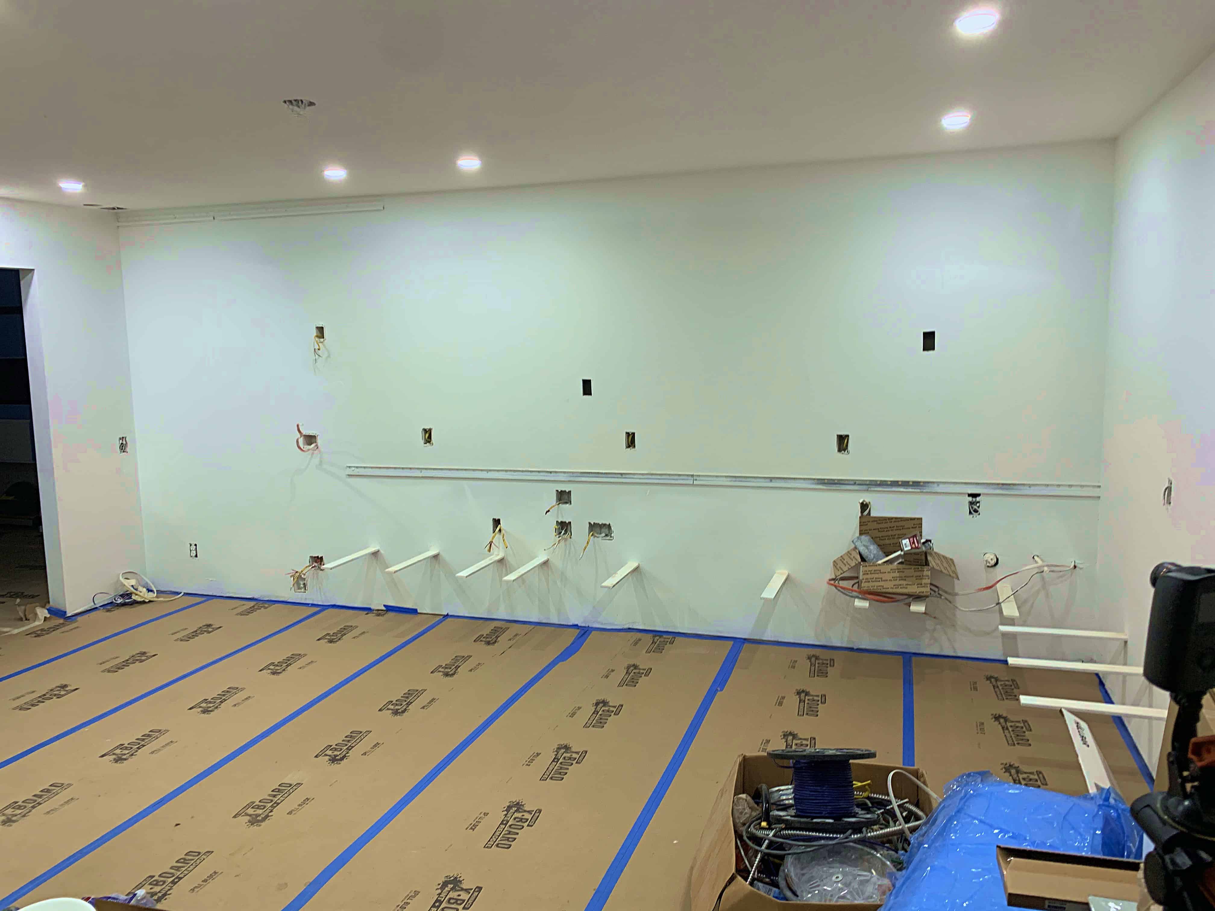 A partially finished room with a light green wall, multiple electrical outlets, and exposed wires. The floor is covered with cardboard protection, and blue painter's tape outlines the edges.