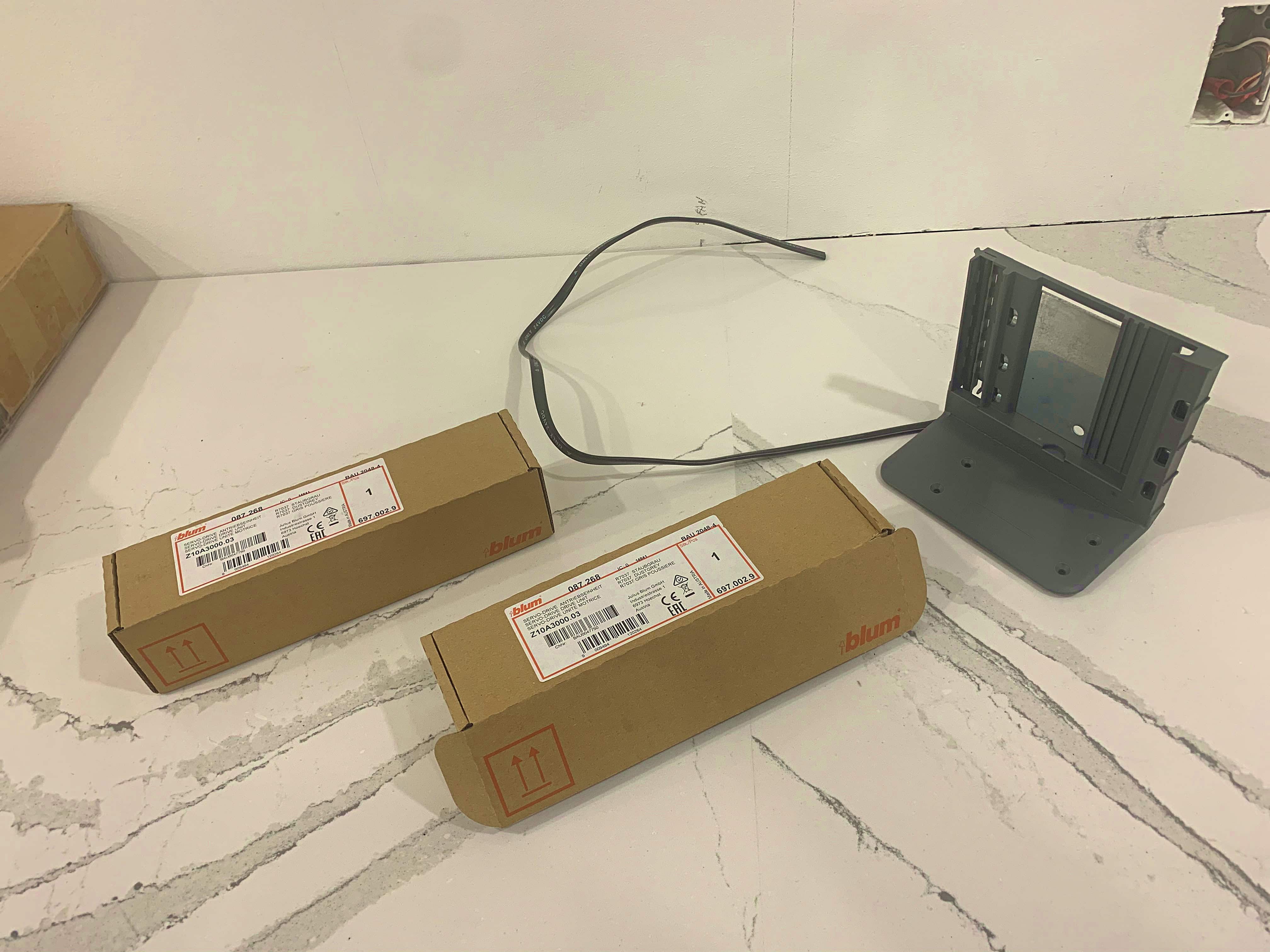 Two brown boxes labeled 'blum' containing hardware parts, alongside a gray mounting bracket and a black cable on a white marble surface.