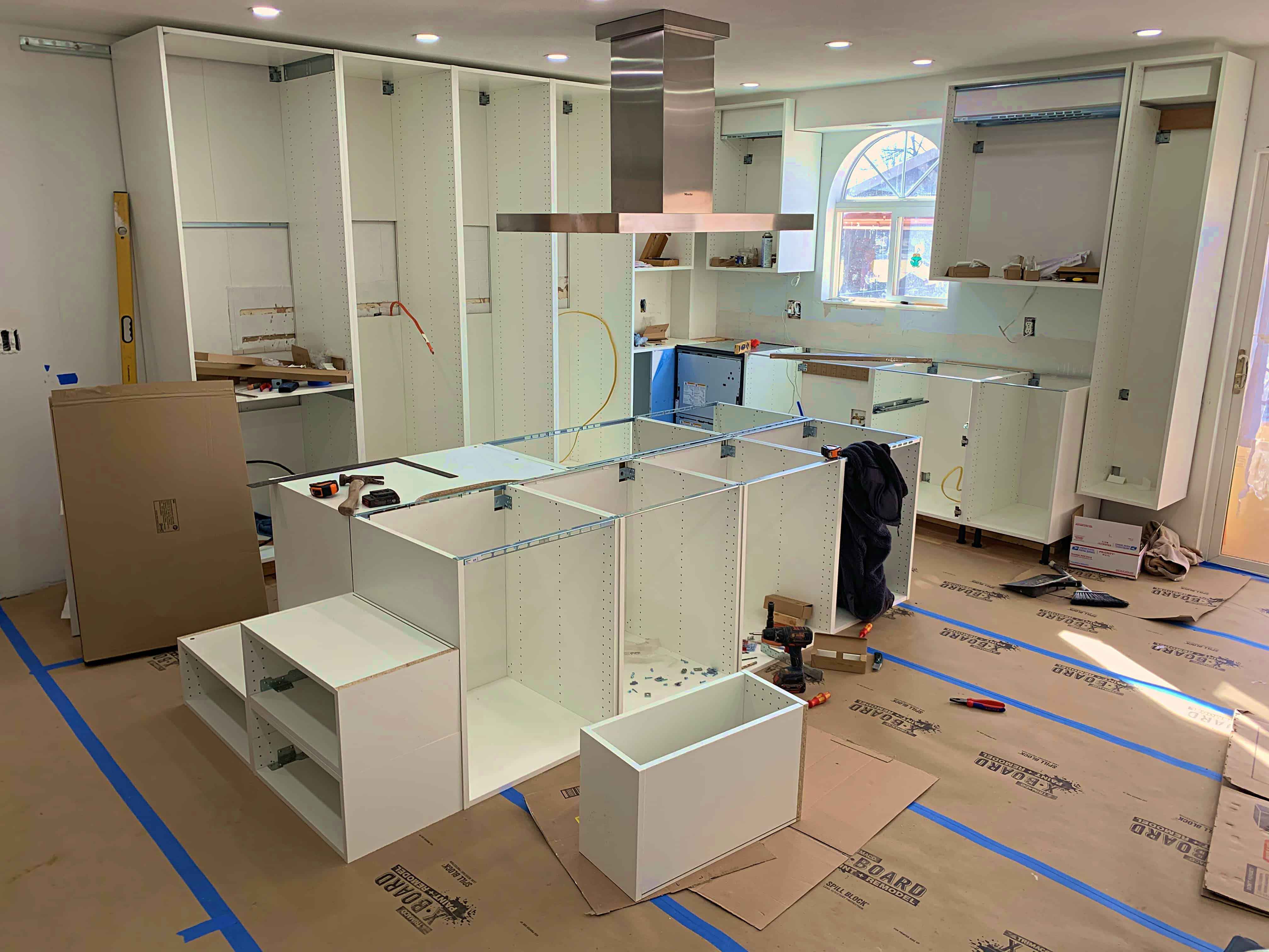 An unfinished kitchen installation with white cabinets and countertops. The space features various cabinet units positioned throughout, tools scattered on the floor, and protective coverings on the ground.