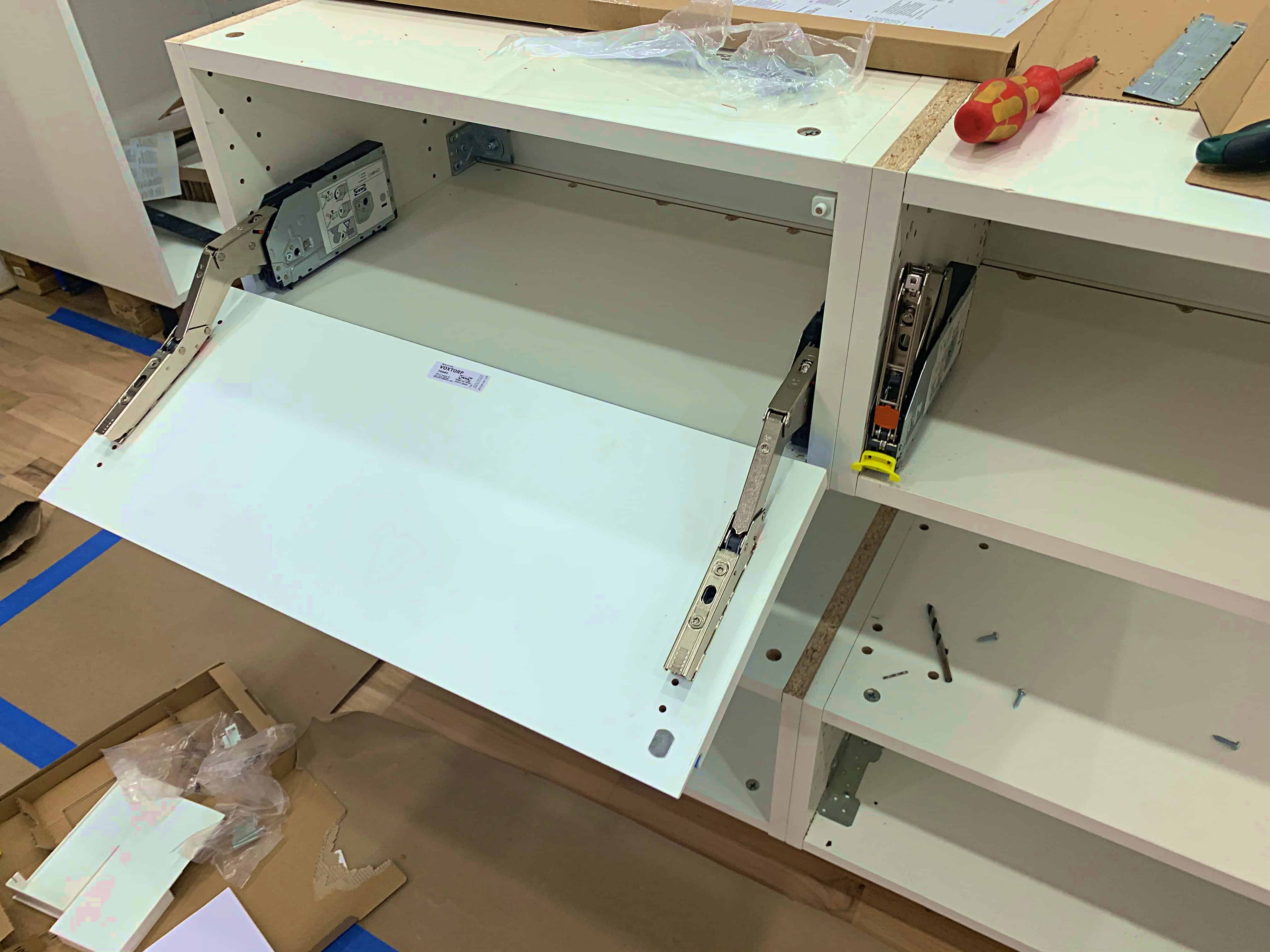 Close-up of an unfinished cabinet with a partially installed drawer slide mechanism. The drawer is open, revealing a white front and the interior cabinet space. Tools and packaging materials are visible nearby.