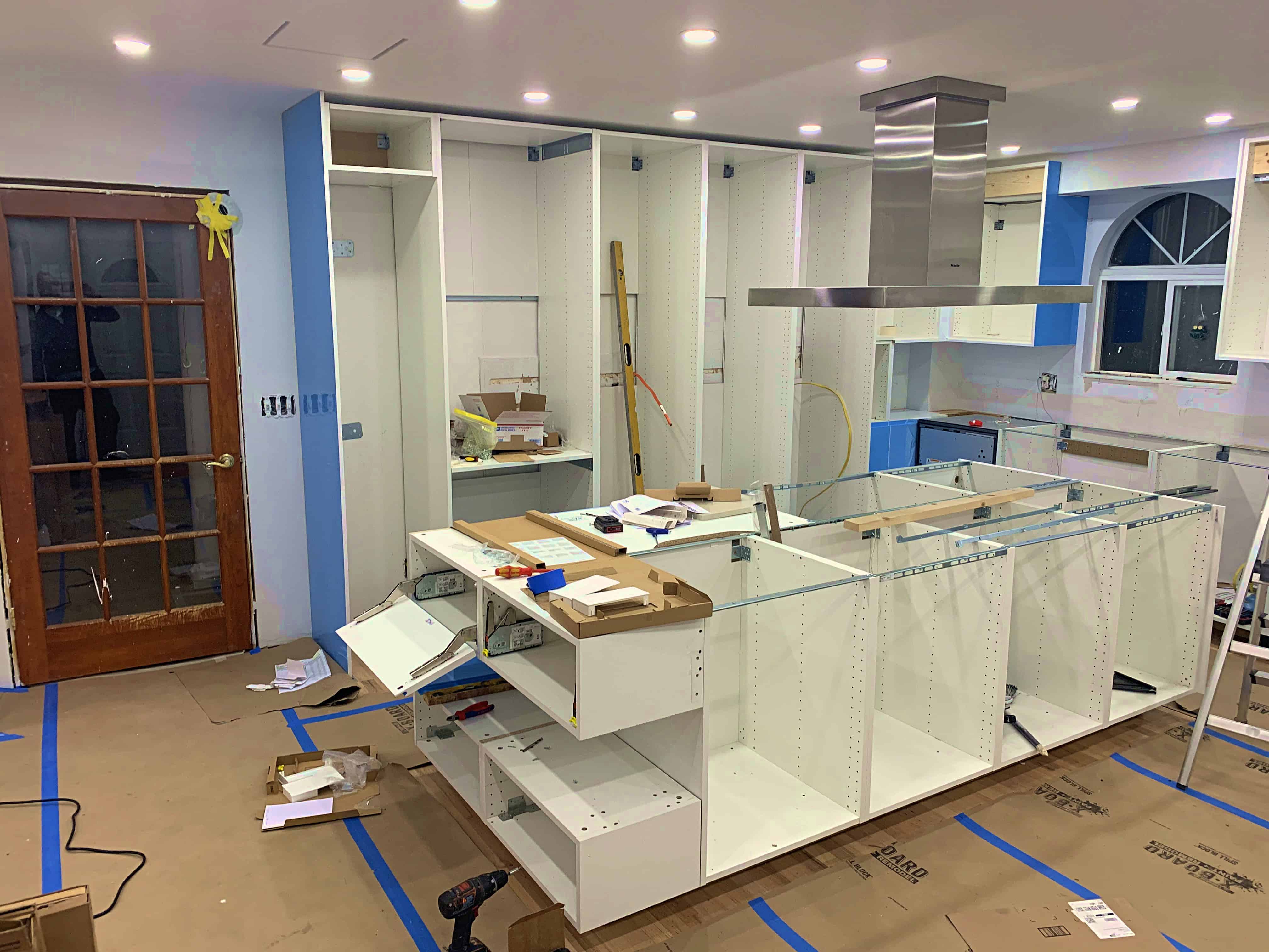 A partially constructed kitchen with white cabinetry and an overhead stainless steel range hood. The floor is covered with cardboard, and tools and materials are scattered throughout the space.