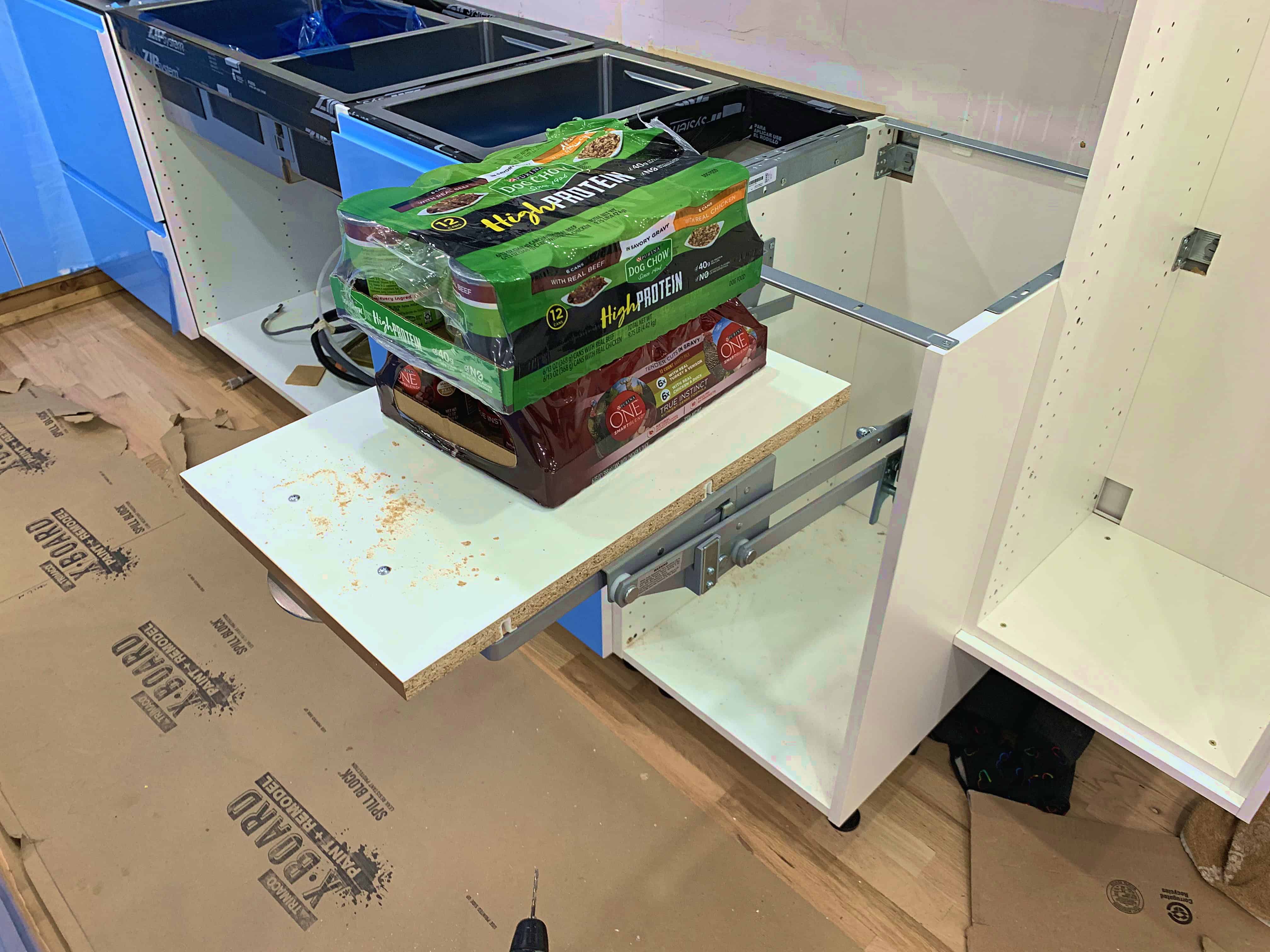 An unfinished kitchen setup showing an empty cabinet with pulled-out drawers and a shelf holding protein snacks, on a wooden floor covered with cardboard.