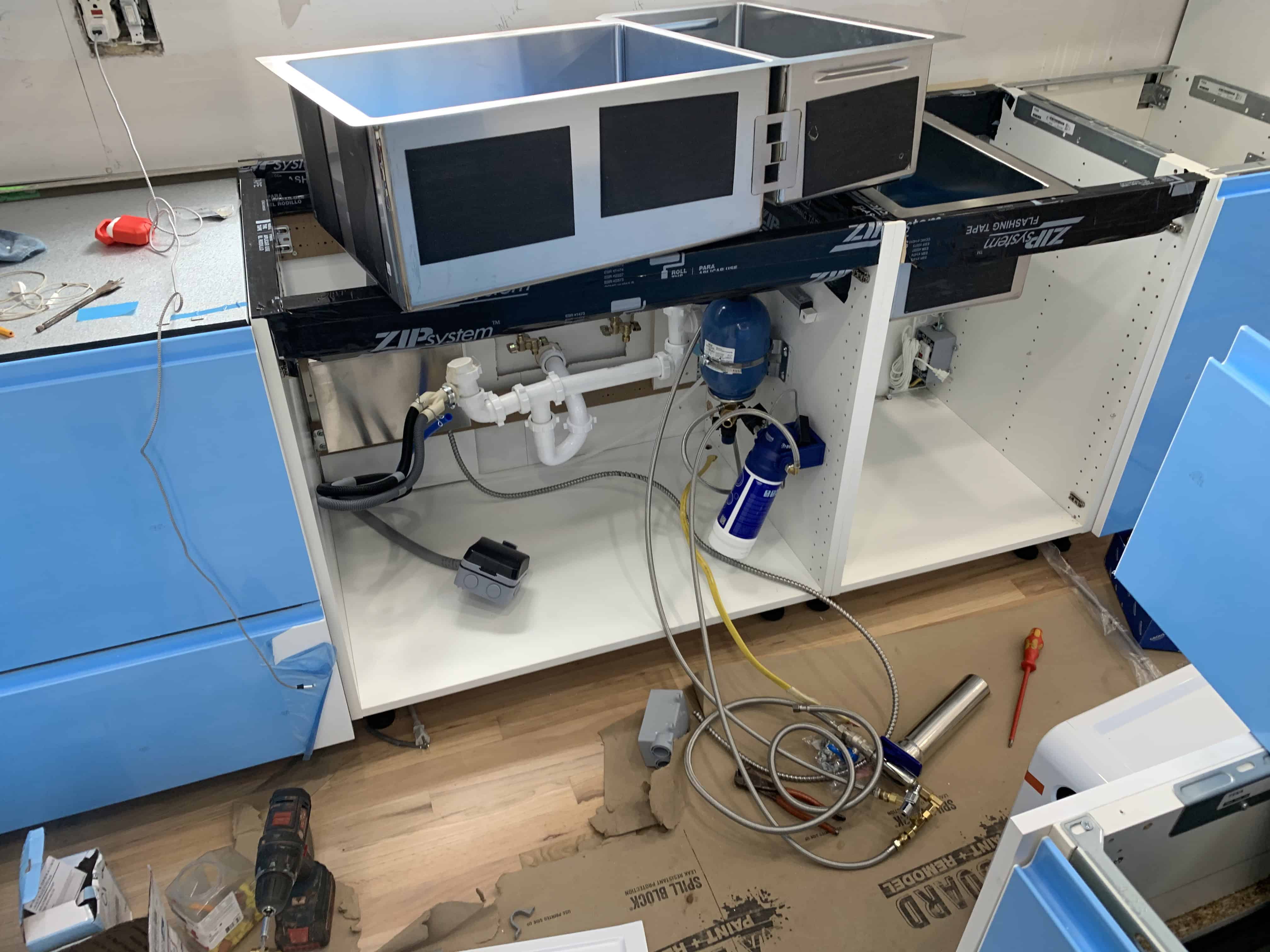 A kitchen cabinet installation in progress, featuring two microwave units, plumbing connections, and various tools scattered on the floor.