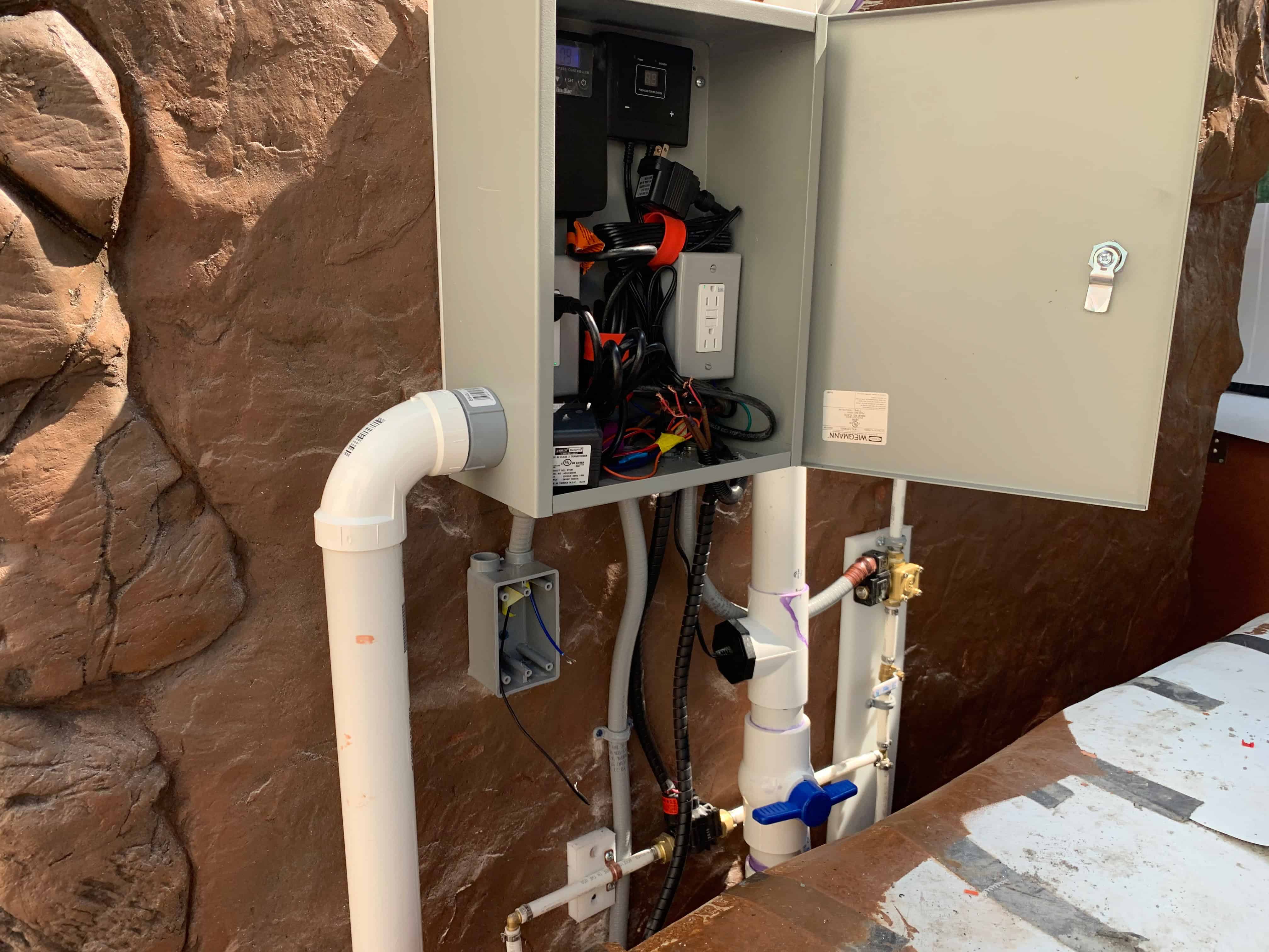 Control panel with pump, filtration, and plumbing mounted on the fountain's rock wall.