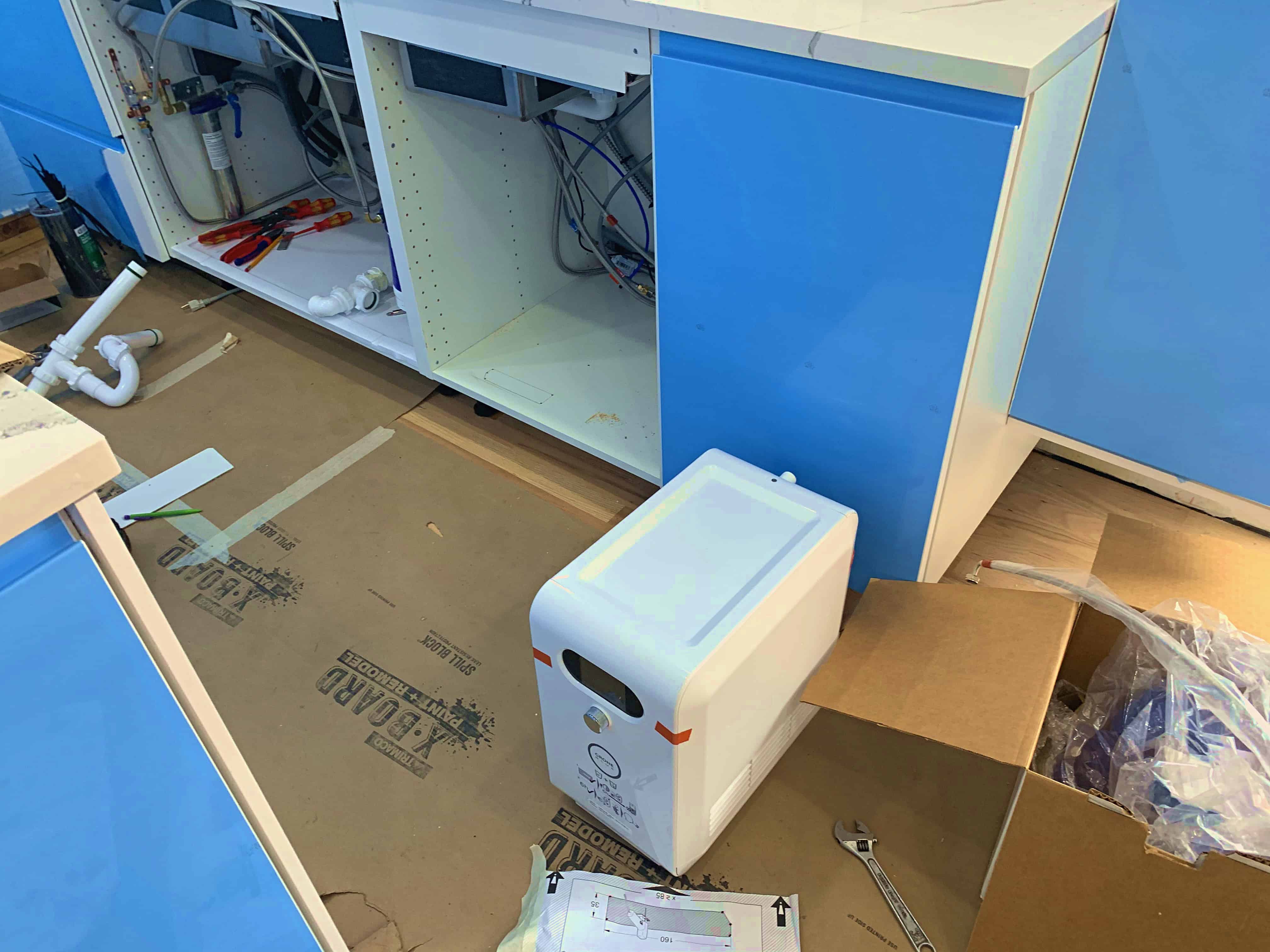 A partially assembled kitchen cabinet with blue doors, revealing plumbing and electrical components inside. A small white appliance is placed on the floor next to cardboard packaging and tools.