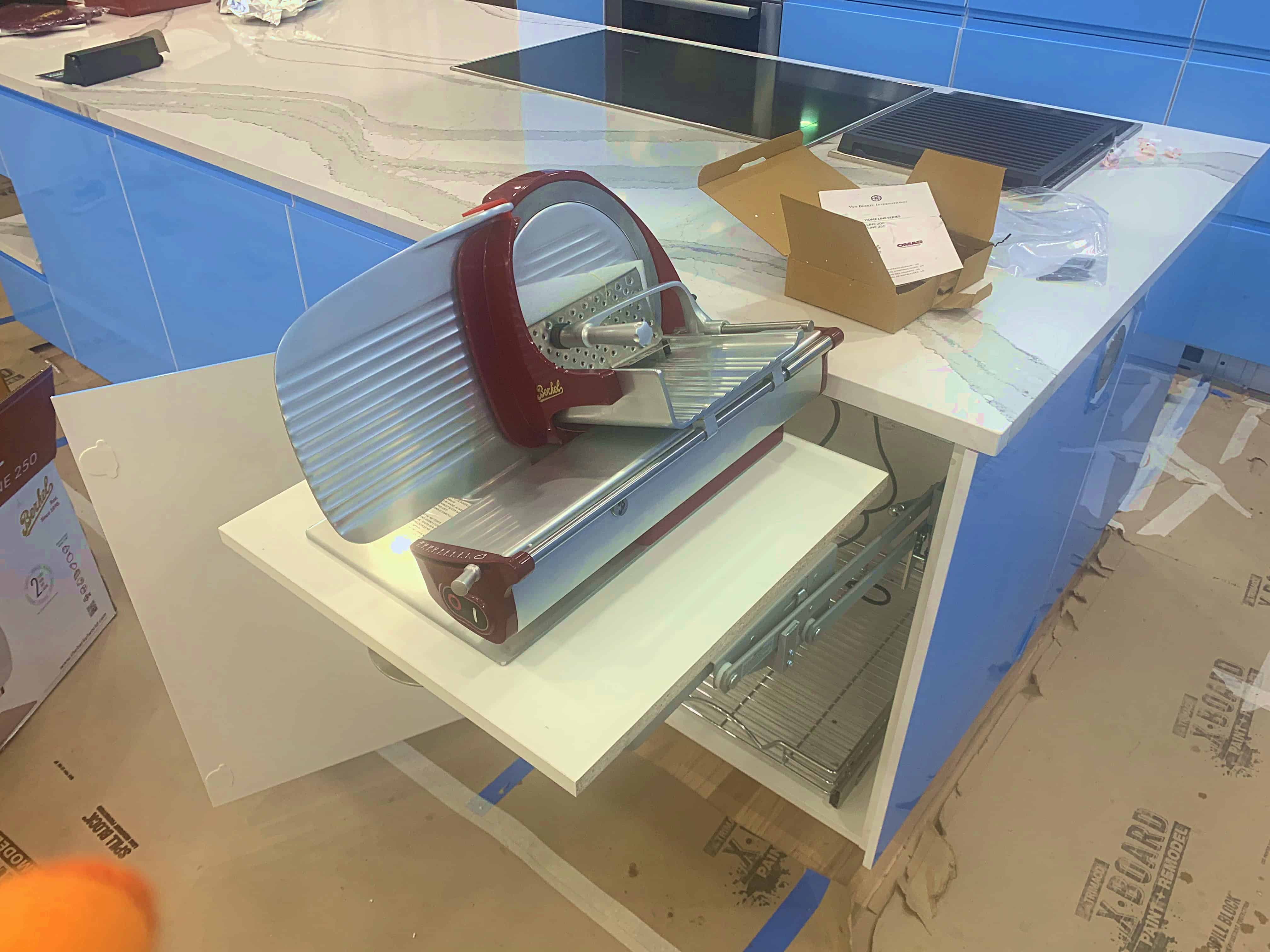 A red and silver meat slicer placed on a countertop with a modern design kitchen background. Boxes and packaging materials are visible nearby.
