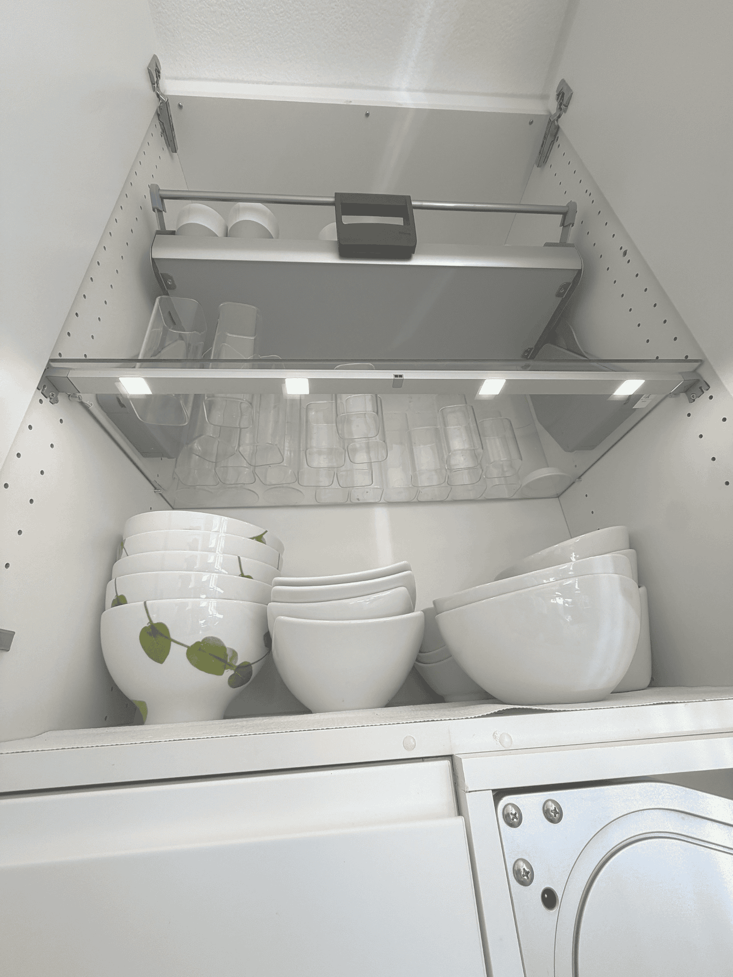 Glass shelf of bowls and glasses lit by an IKEA MITTLED sensor light, above the iMove pull-down shelf in a 24-inch SEKTION cabinet