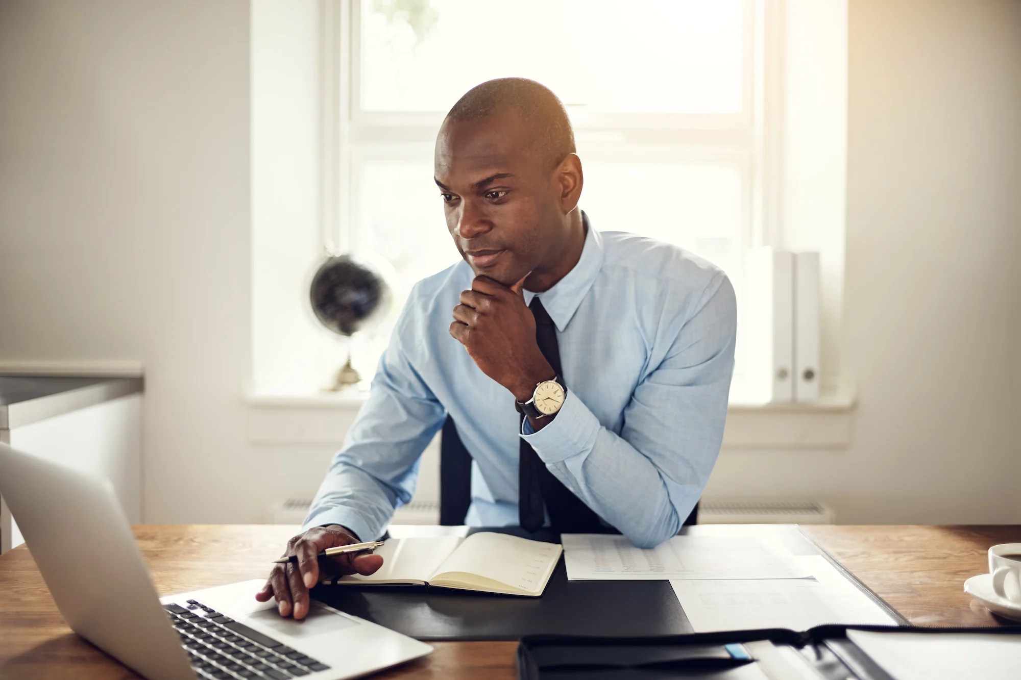 A private investigator in a blue shirt and tie sits at a desk with a laptop, notebook, and papers, looking thoughtfully at the screen.