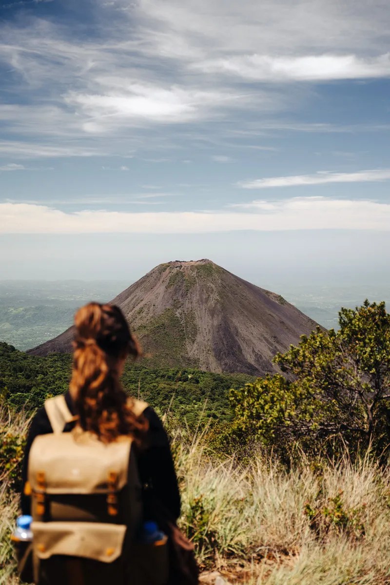 Turista con mochila contemplando el majestuoso cono del Volcán de Izalco desde un sendero rodeado de naturaleza en El Salvador.