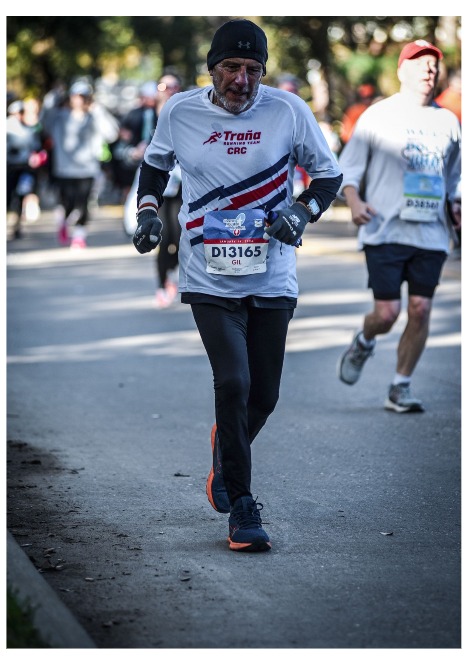 Un corredor mayor participando en una carrera, vestido con una camiseta de color blanco y pantalones negros, mientras otros corredores lo acompañan en el fondo.