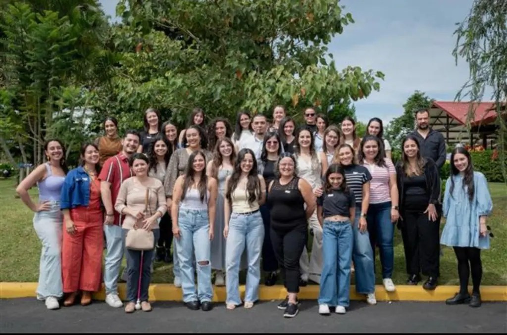 Grupo de personas posando juntas al aire libre, con un fondo de árboles y un edificio. Todas parecen estar disfrutando del momento.