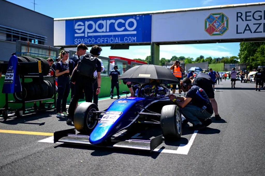 Auto de carrera azul estacionado en la pista, con varios mecánicos y miembros del equipo alrededor, preparándolo para la competencia.