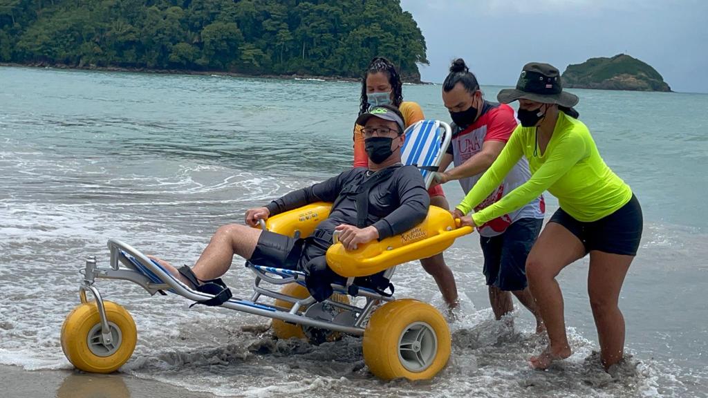 Personas ayudando a una persona en una silla de ruedas acuática en la playa, con mar y vegetación de fondo.