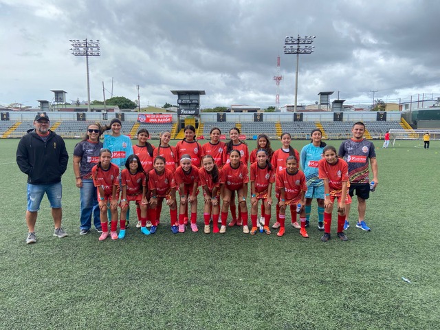 Equipo de fútbol femenino posando en el campo con uniforme rojo y pantalones cortos. Hay entrenadores y un grupo de jugadoras sonriendo al frente. El fondo muestra gradas vacías y un cielo nublado.