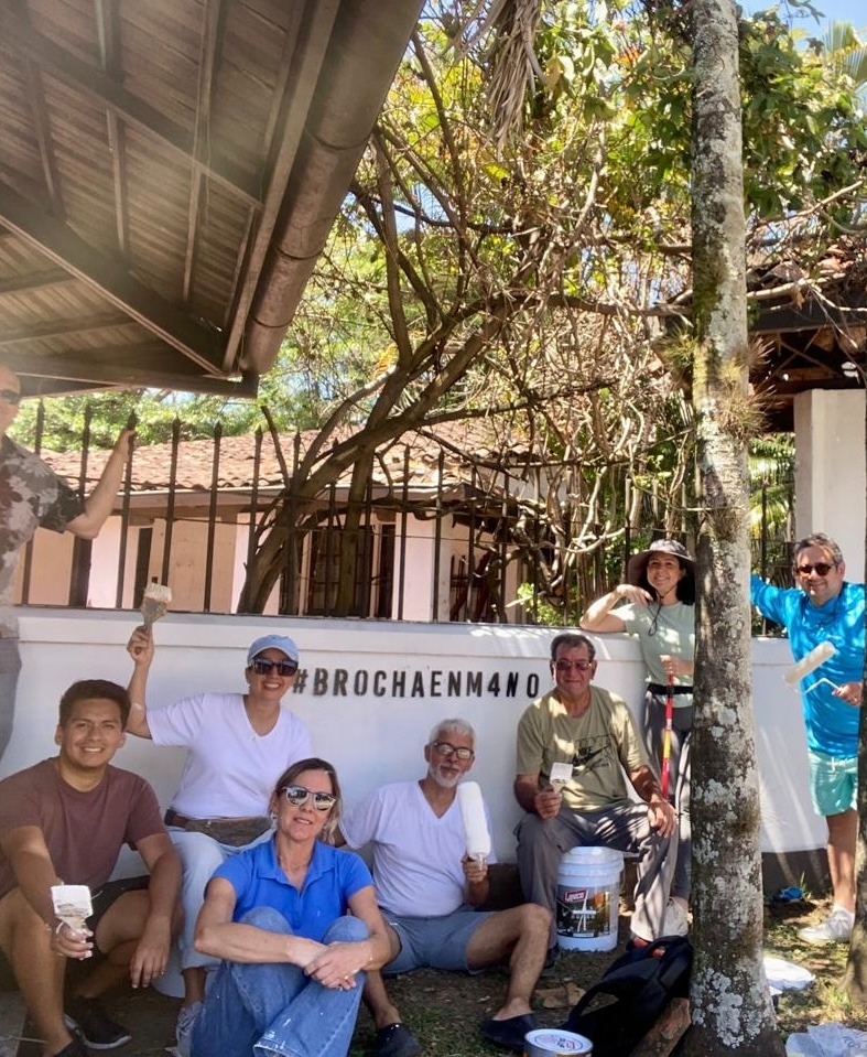 Grupo de personas posando con pinceles y rodillos de pintura frente a un muro con el hashtag #BROCHAENM4O, en un ambiente al aire libre.