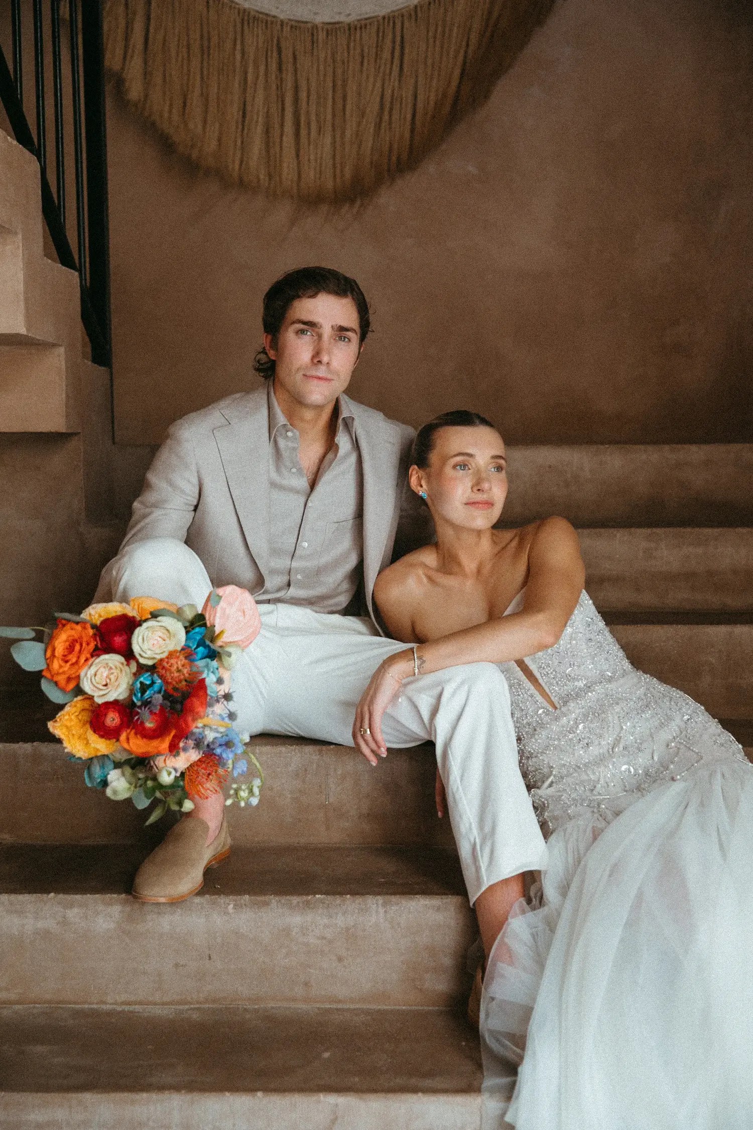 Laura and Stephen sit on a terracotta staircase with a dense orange, red and blue bouquet in the middle.