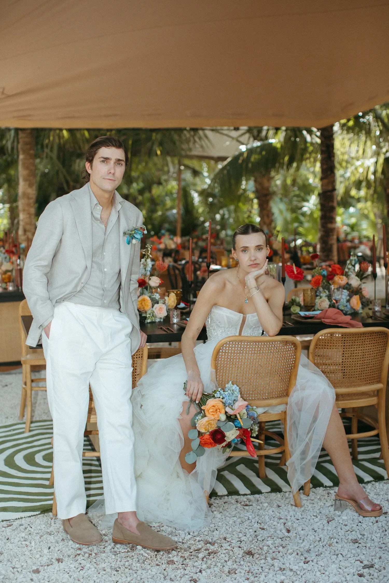 Laura sits with her bouquet and Stephen leans on the long reception table under the canvas tent.