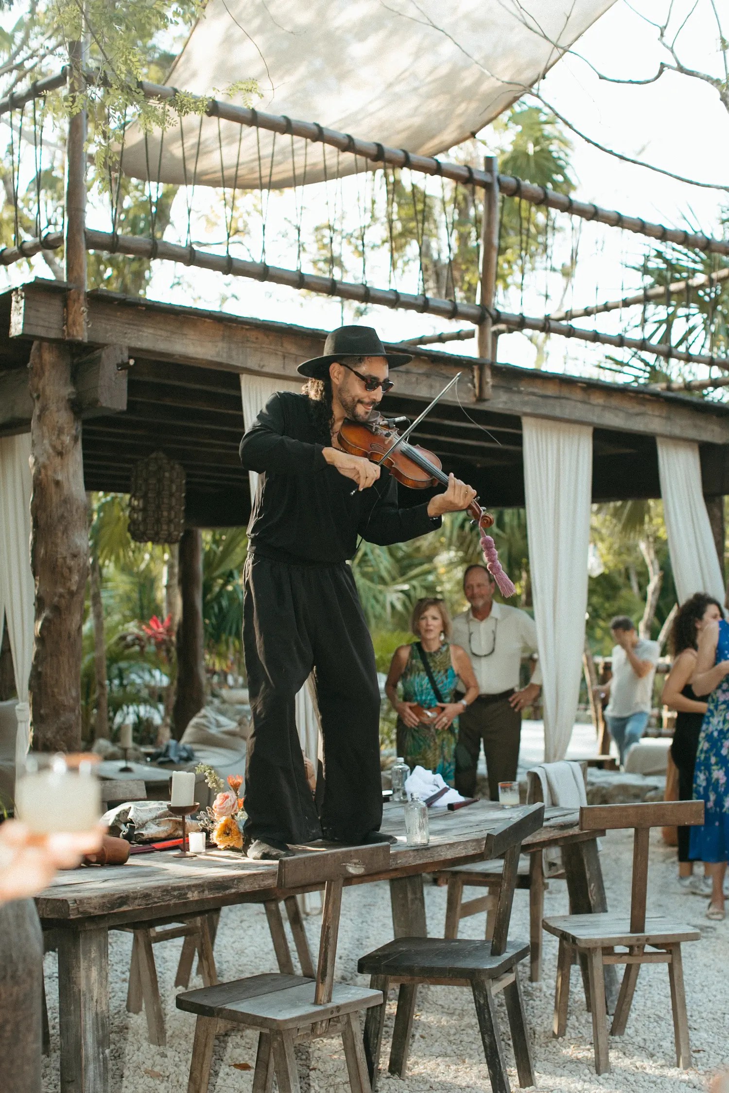 Diego the violinist dressed in all black sports standing at a rustic wooden table at an outdoor reception.