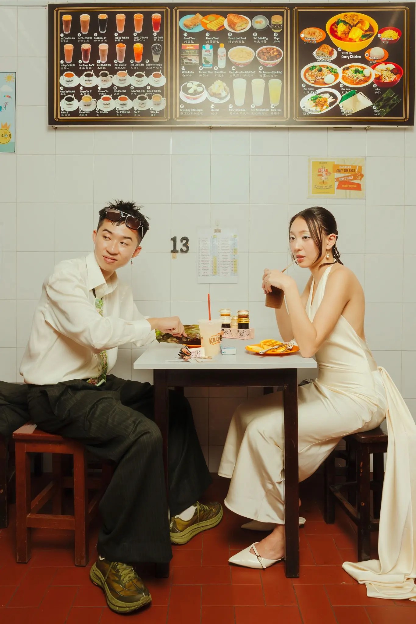 Couple assis à une table kopitiam avec des toasts kaya et du café glacé sous un tableau de menu photo pleine largeur, Rachel dans une robe dos nu ivoire