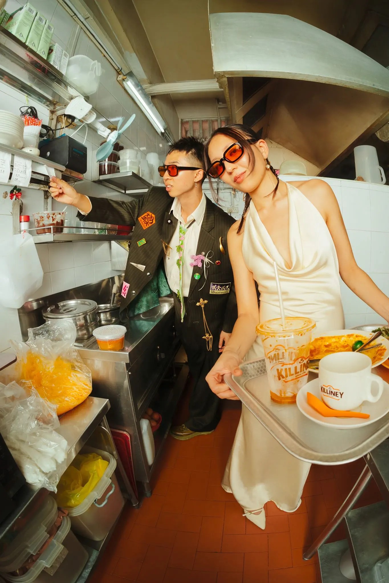 Couple portant des lunettes de soleil ovales rouges à l'intérieur d'une cuisine kopitiam, Rachel tenant un plateau Killiney avec une boisson glacée, une tasse de café et du pain grillé kaya