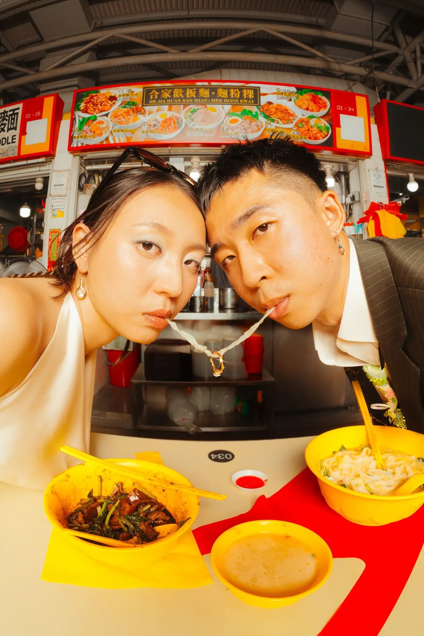 Rio et Rachel partagent un brin de nouilles sur une table de vendeur ambulant avec des bols jaunes et un plateau 034, photographiés en fish-eye depuis le niveau de la table.