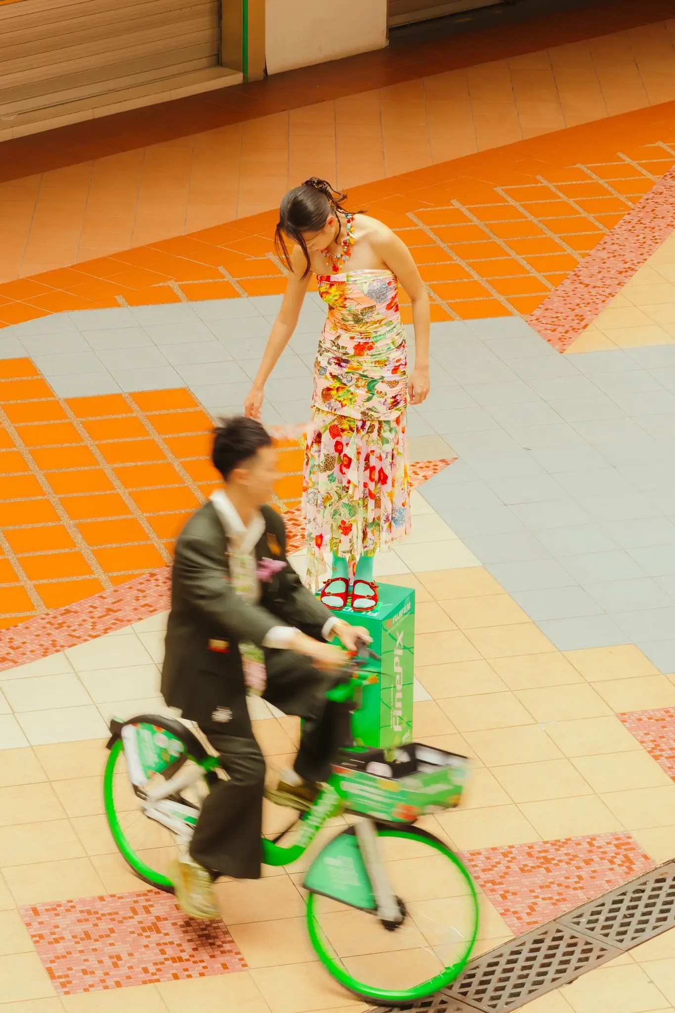 Rio sur un vélo partagé vert dans un flou de mouvement sur une terrasse vide de Singapour en mosaïque alors que Rachel en robe à fleurs tend la main vers le vélo d'en haut