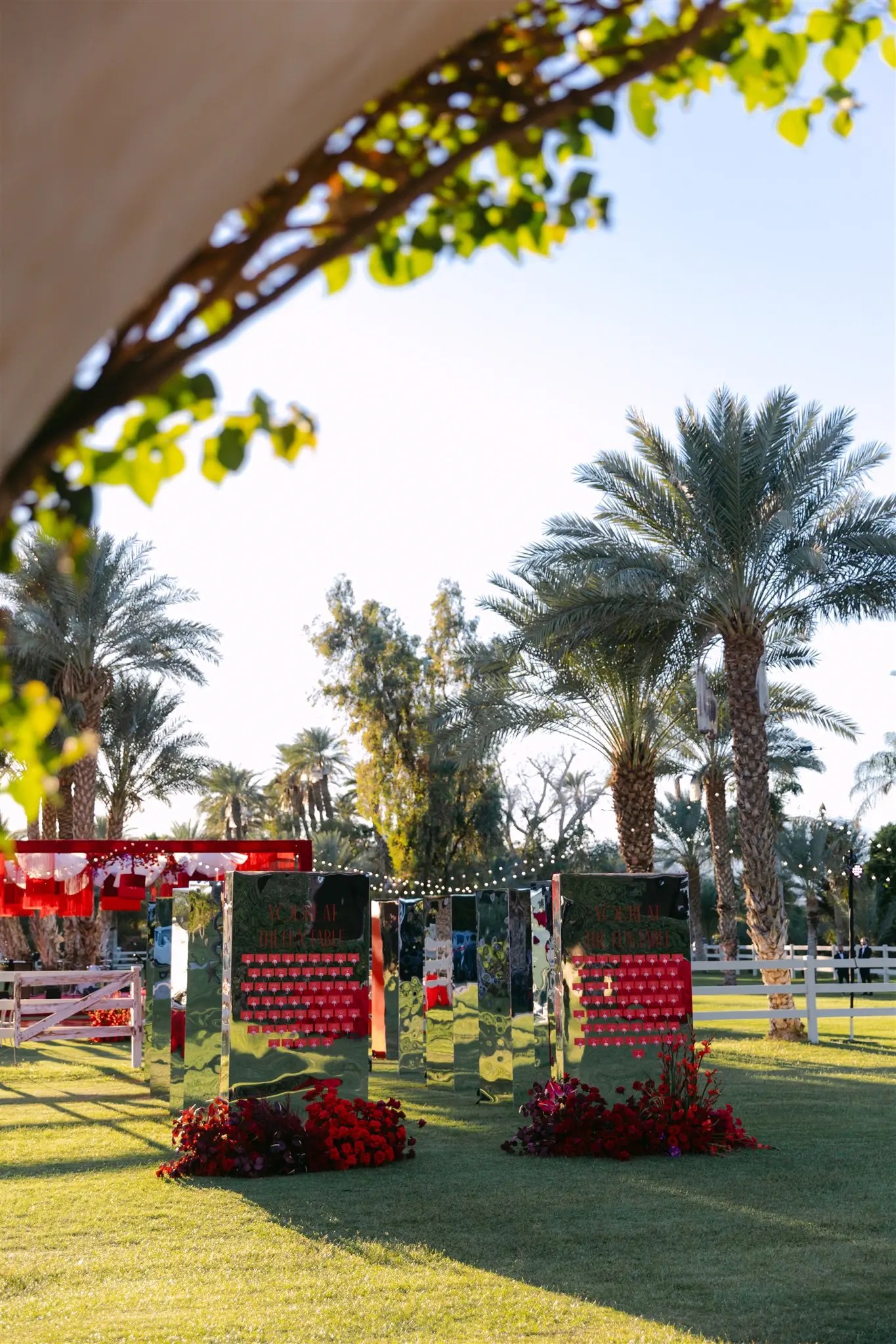 Three tall chrome mirror seating chart panels covered in red escort cards with low red and burgundy flower arrangements at the feet and globe string lights between them on the estate lawn in the evening.